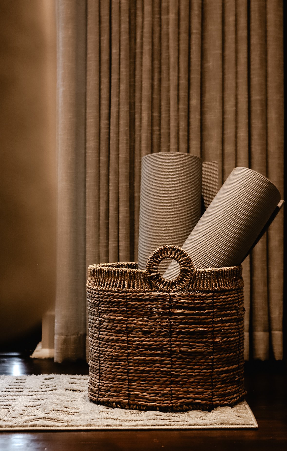 A woven basket holding three rolled yoga mats is placed on a rug in front of brown curtains.