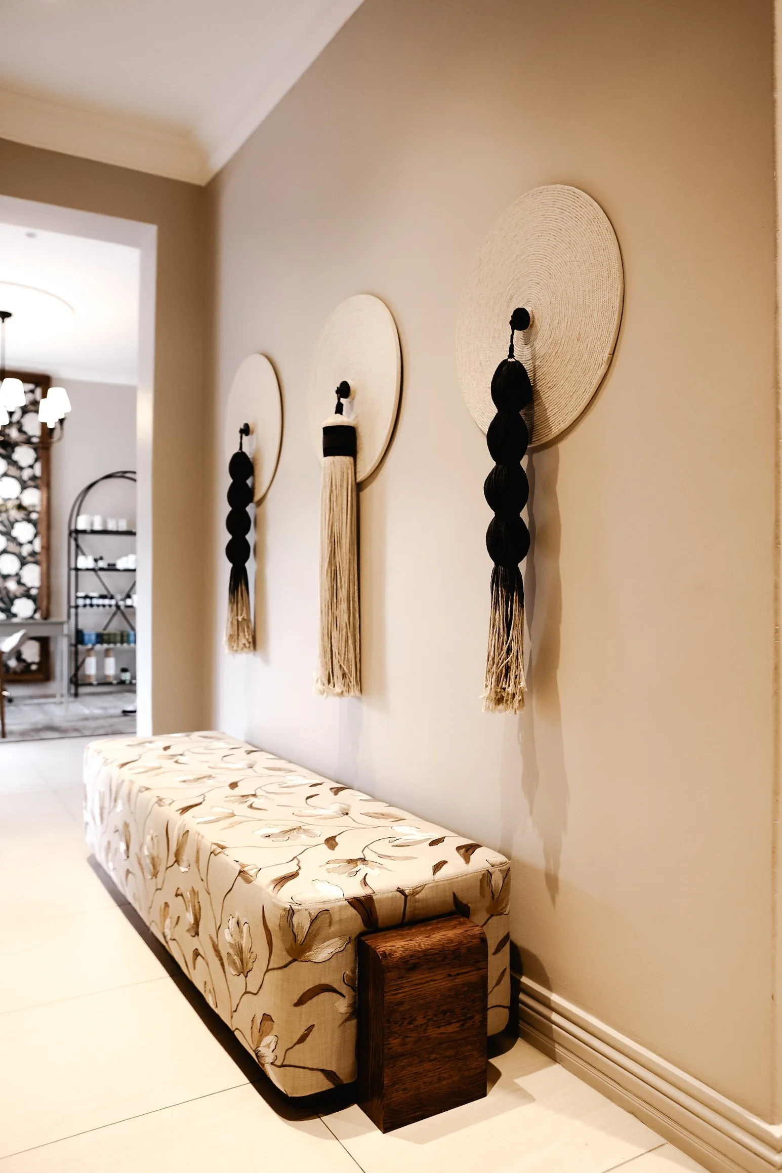 A beige floral-patterned cushioned bench next to a dark wood side table against a plain wall, with three decorative wall hangings with long black and beige tassels hanging below round woven discs.