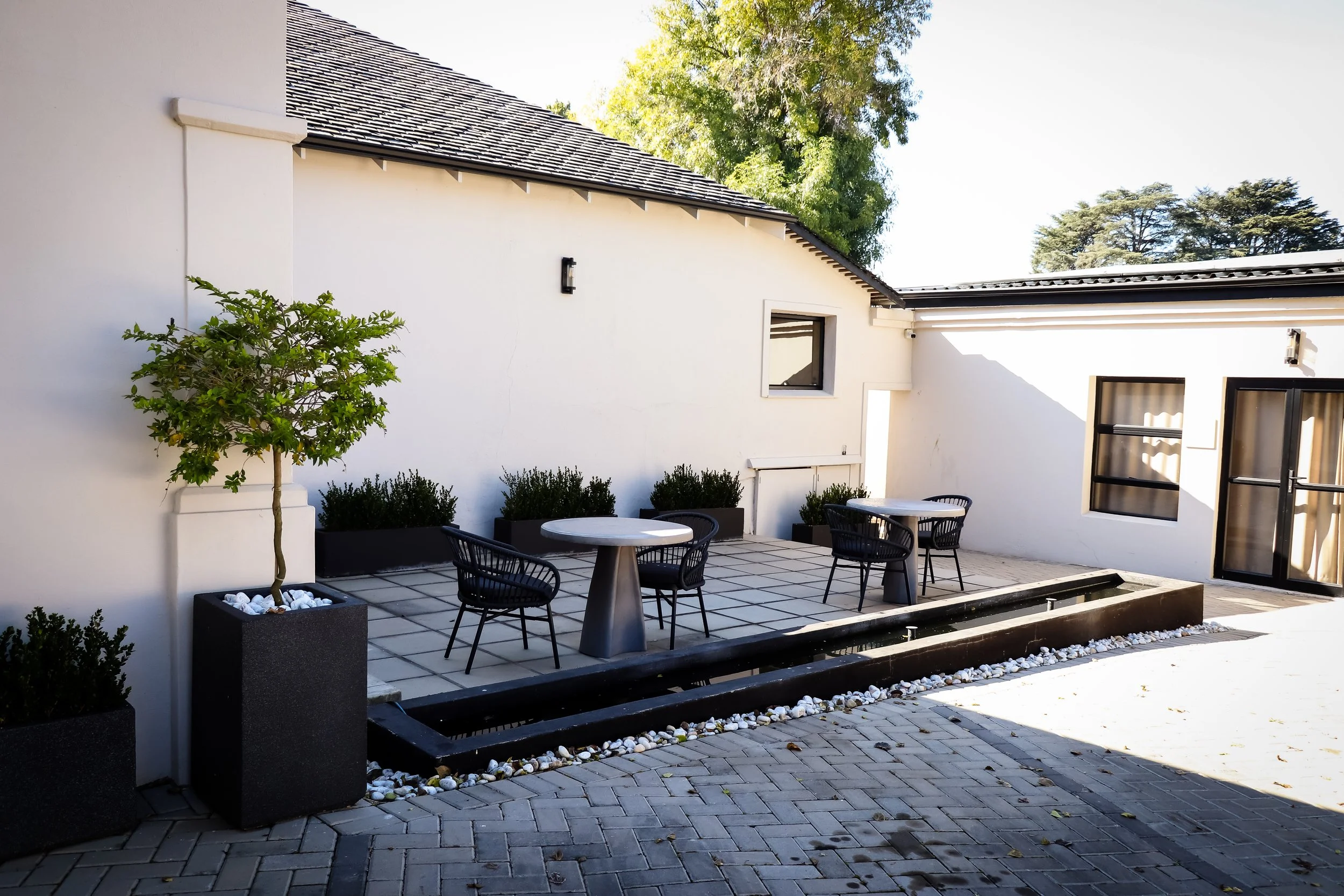 A modern outdoor patio with black chairs and round tables, potted greenery, and a white house exterior with windows and a door, surrounded by trees.