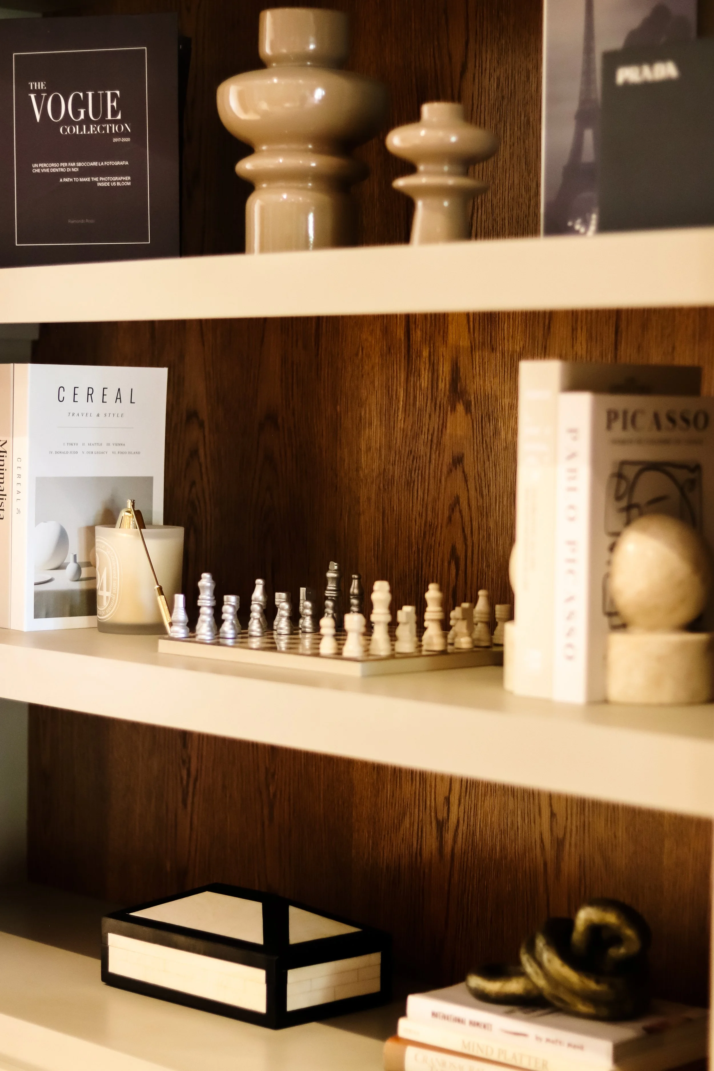 Bookshelves featuring decorative items, including two ceramic vases, a chess set, a white candle, and various fashion and art books like Vogue Collection, Picasso, and a book titled "Cereal".