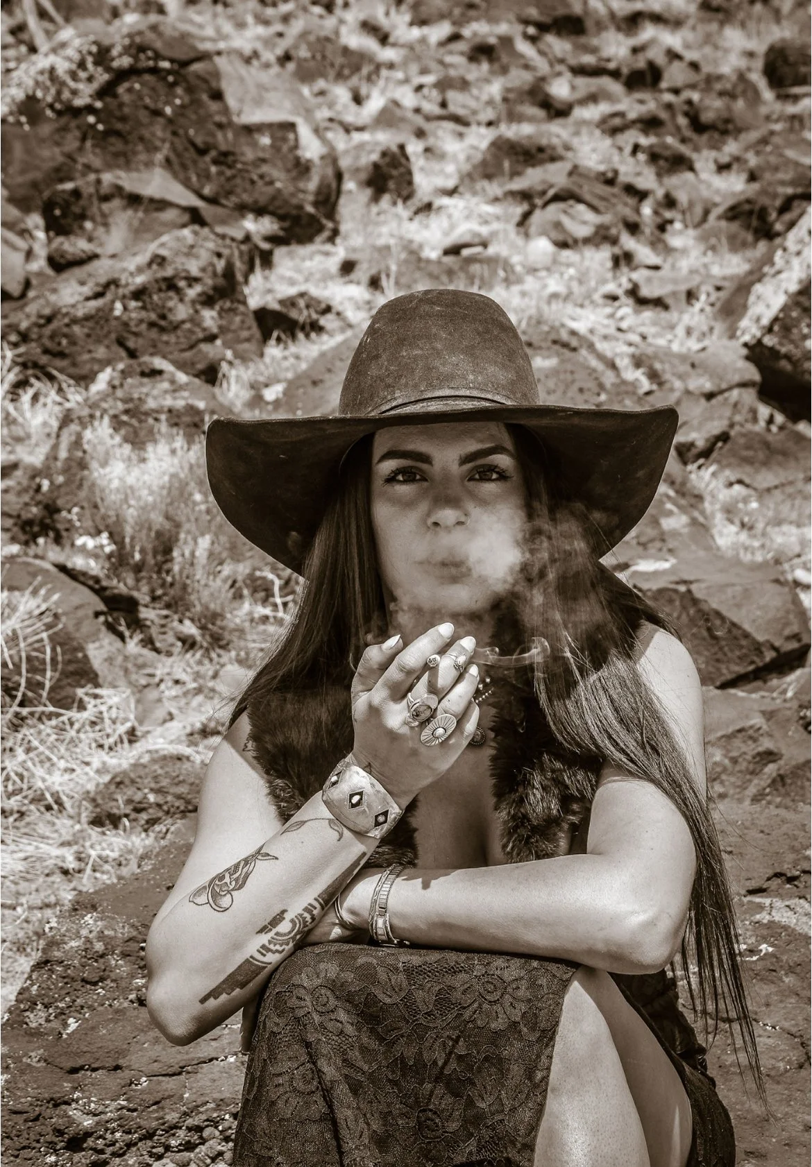 A woman wearing a large wide-brimmed hat, sleeveless dress, and jewelry sits outdoors on rocky terrain, smoking a cigarette.