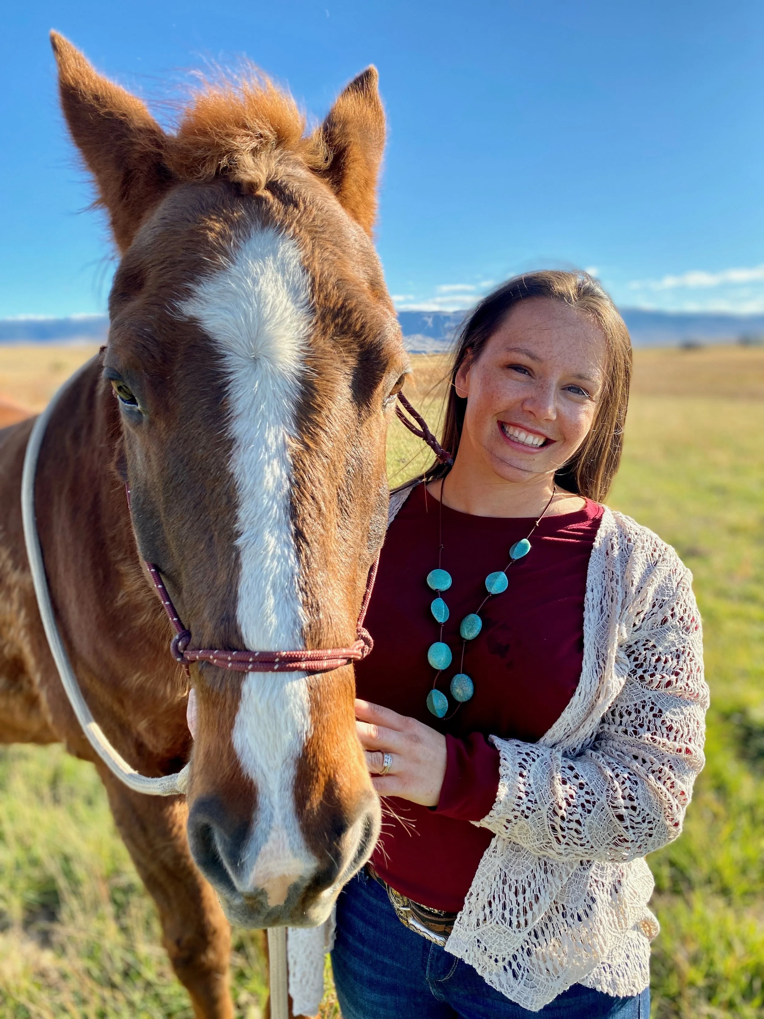 A woman smiling and holding a brown and white horse in a field with mountains in the background under a clear blue sky.