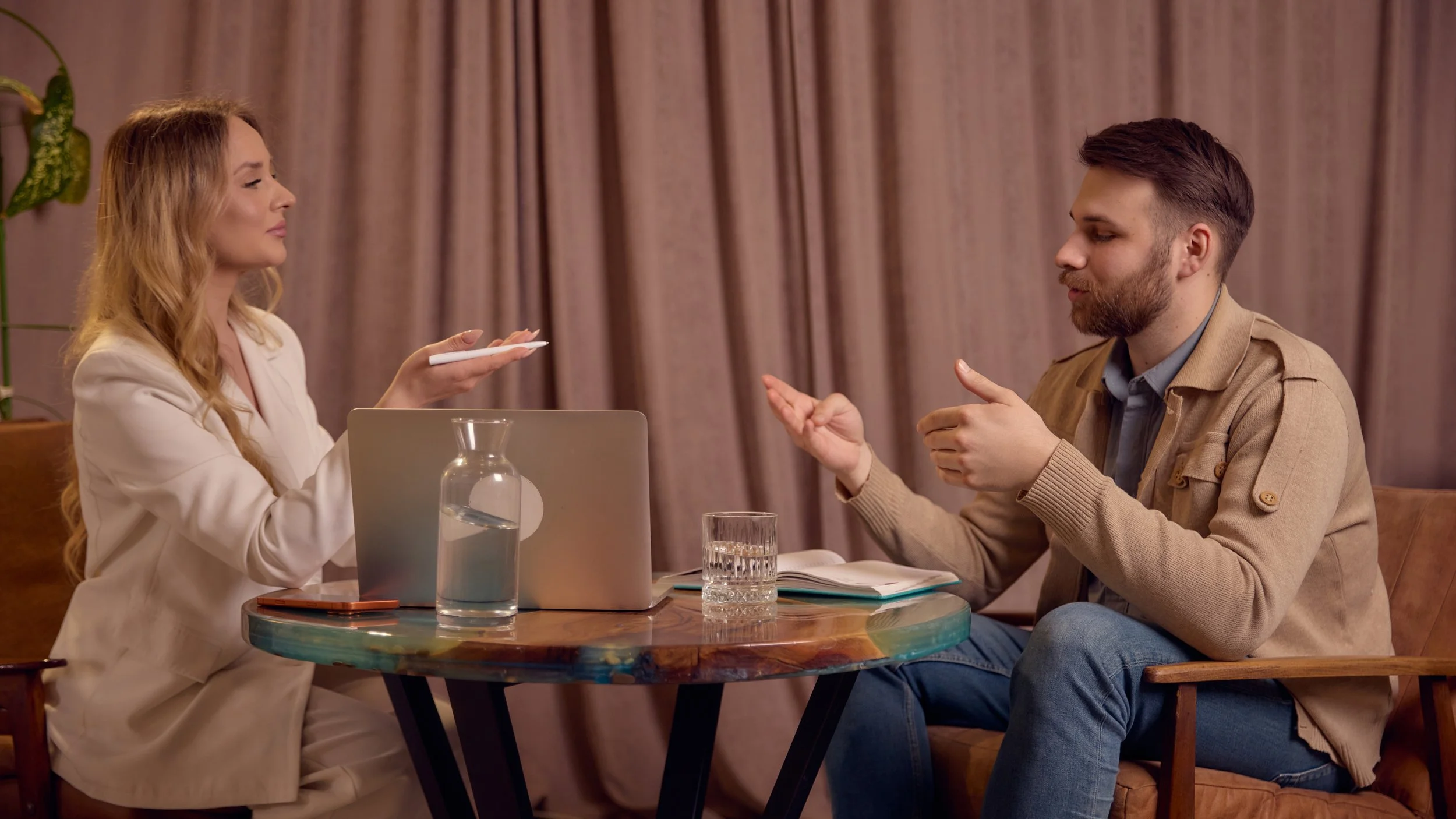 A woman and a man engaging in a conversation at a table. The woman has long, wavy blonde hair and is wearing a beige blazer. She has a laptop in front of her, along with a glass of water and a vase with water. The man has short brown hair, a beard, and is wearing a beige jacket over a blue shirt. They are sitting in a room with curtains in the background.