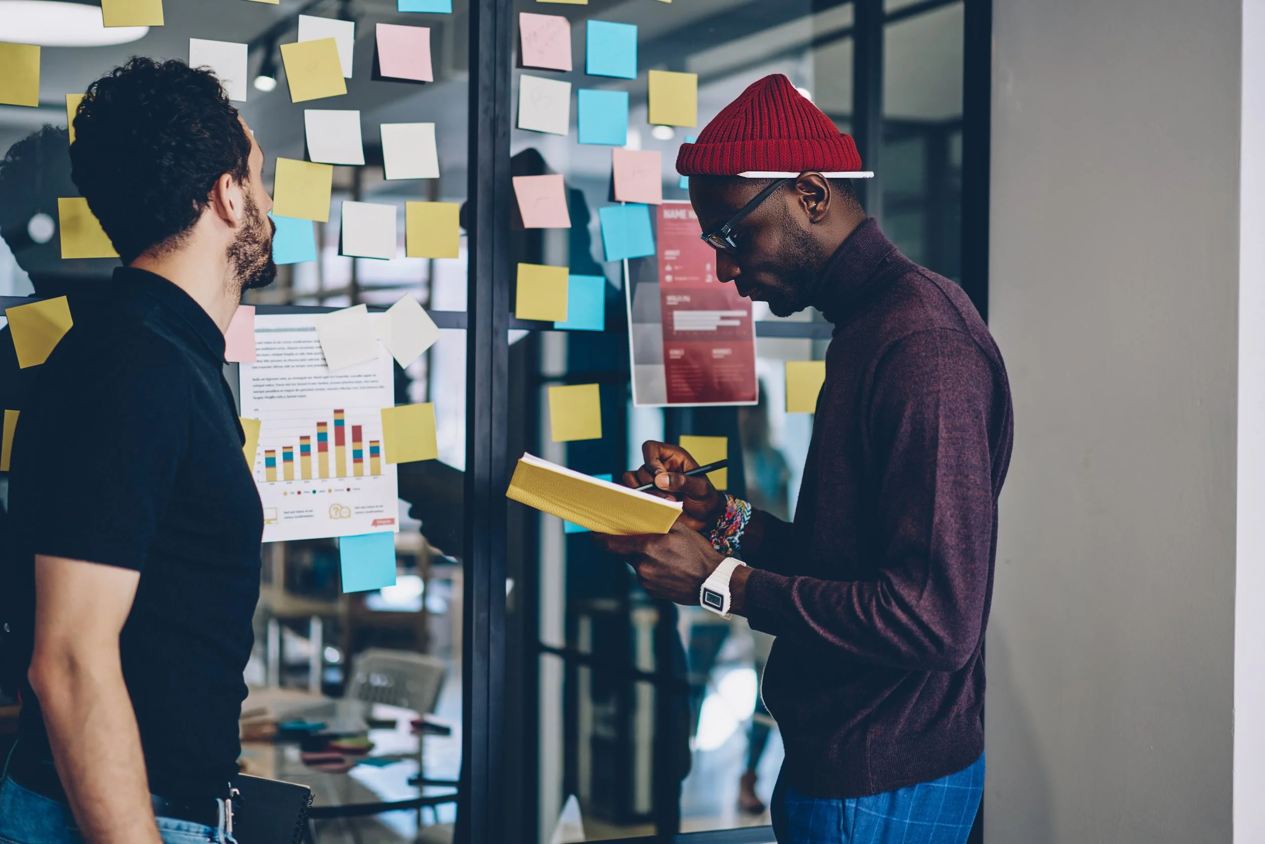 Two men are standing in an office space with a wall of colorful sticky notes and printed charts. One man is holding a yellow folder and a pen while the other man is looking at him, wearing a red beanie, glasses, and a smartwatch.