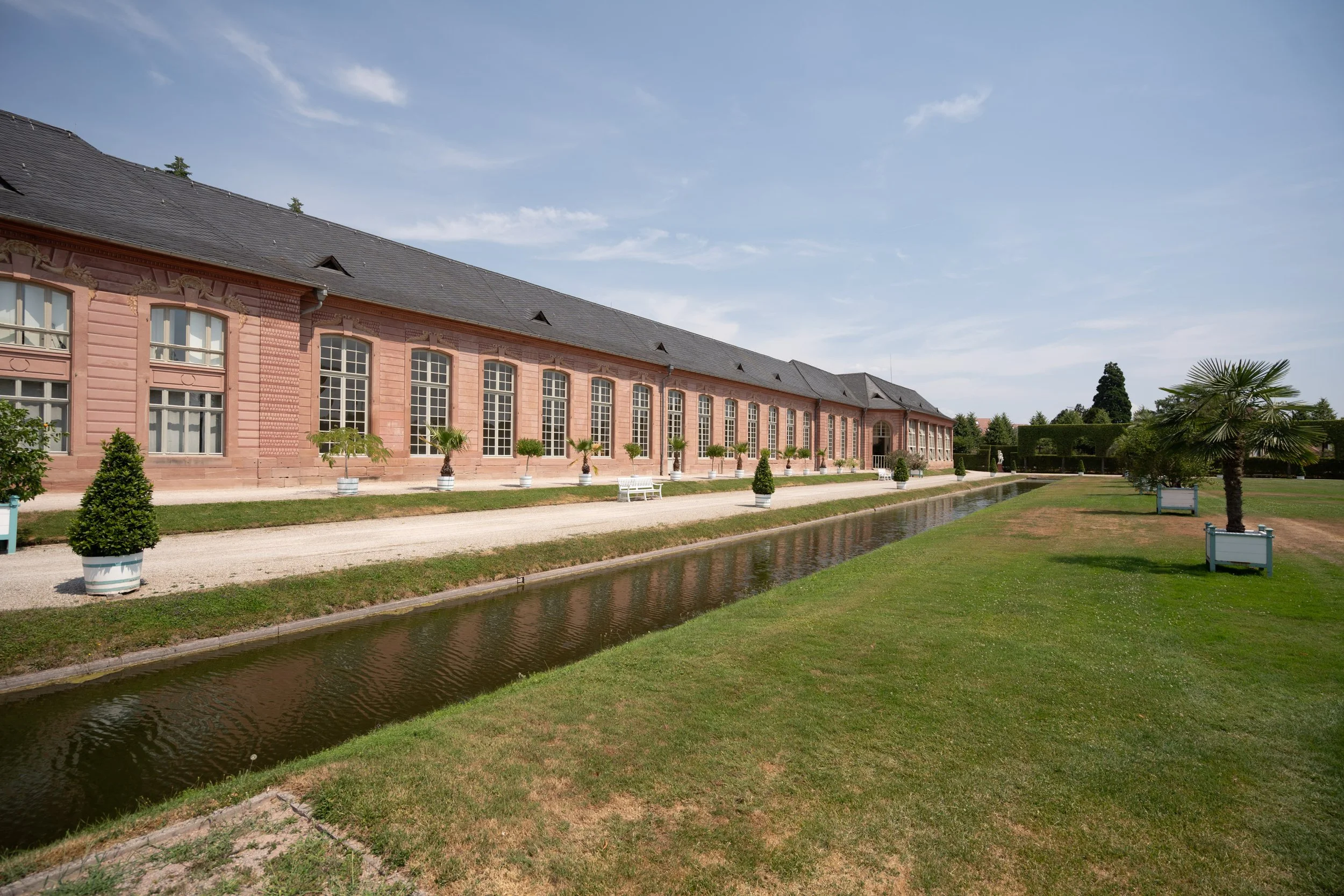 Ein langes, rosa Gebäude mit hohen Fenstern und einem Schieferdach, umgeben von gepflegtem Garten mit Zierbäumen und einem Wassergraben im Vordergrund bei schönem Wetter.