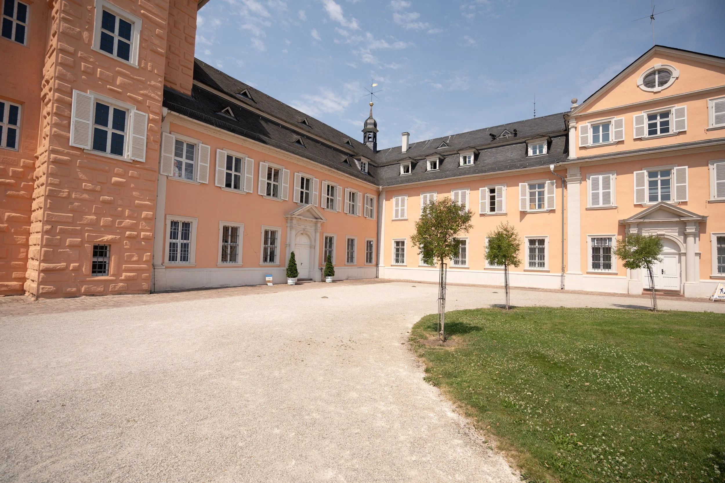 Ein historisches Gebäude mit hellrosa Wänden, weißen Fensterläden und einem schwarzen Dach. Draußen sind kleine Bäume und ein offener Platz mit Kiesboden.