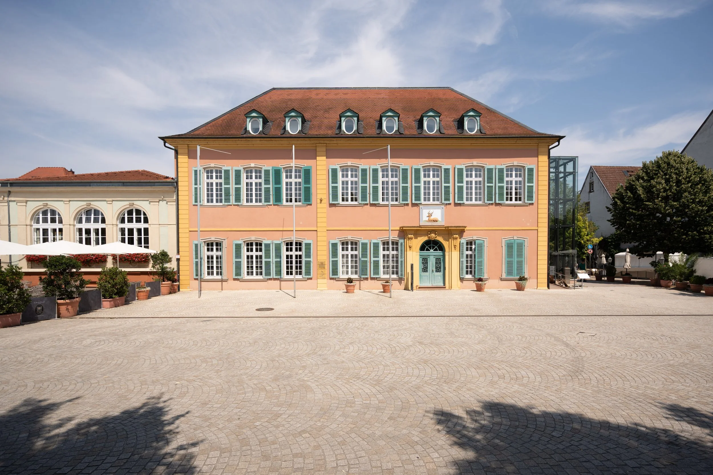 Ein farbiges Gebäude mit mehreren Fenstern und grünen Fensterläden, im Vordergrund eine gepflasterte Plaza mit Schatten von Bäumen und einigen kleinen Pflanzen in Töpfen.