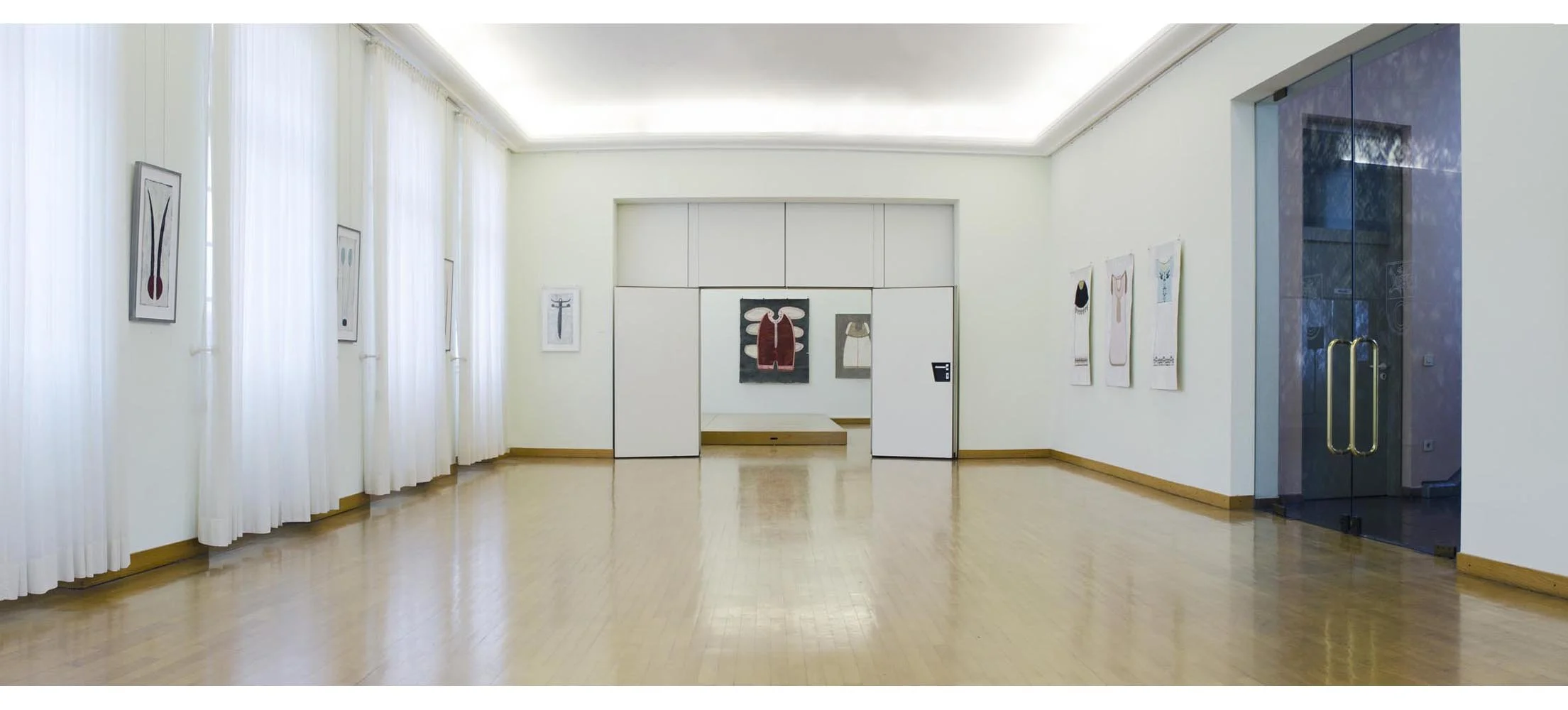 Ein hell erleuchteter Kunstgalerie-Raum mit weißen Wänden und Holzfußboden, der Gemälde und Kunstwerke zeigt, inklusive eines im Zentrum und einen rechteckigen Raum im Hintergrund, mit Glasschiebetüren auf der rechten Seite.
