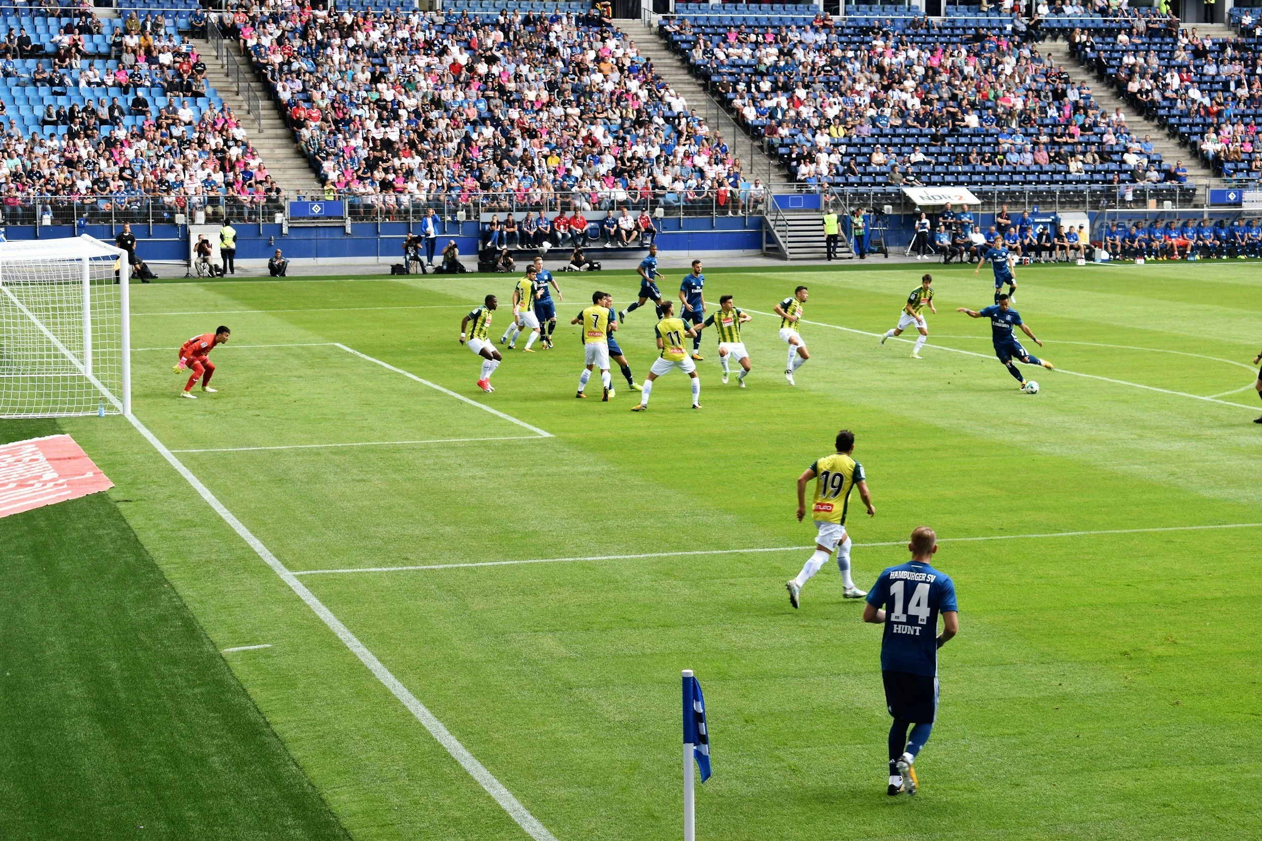 A soccer game in progress on a green field with players from two teams, one in blue jerseys and the other in yellow and black striped jerseys, with a large crowd in the stands. A player in a blue jersey is preparing to kick the ball near the goal, while players in yellow and black are attempting to defend. The audience is seated in rows in the background, and there are officials and photographers along the sidelines.