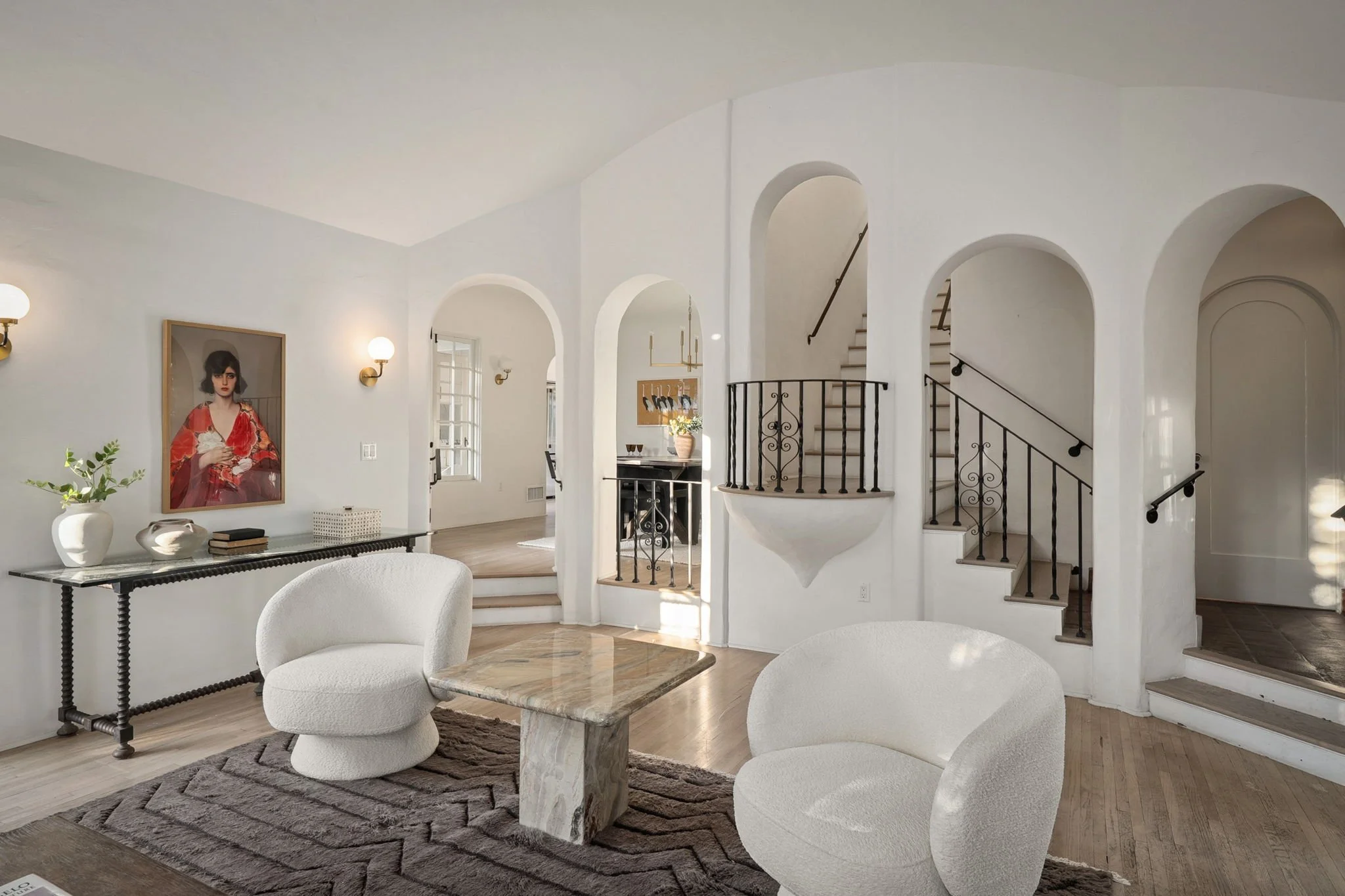 Bright living room with white walls, modern curved white chairs, a marble coffee table, a black console with decorative items, a large brown rug, wall-mounted light fixtures, an art portrait of a woman, an arched staircase with black railing, and side tables.