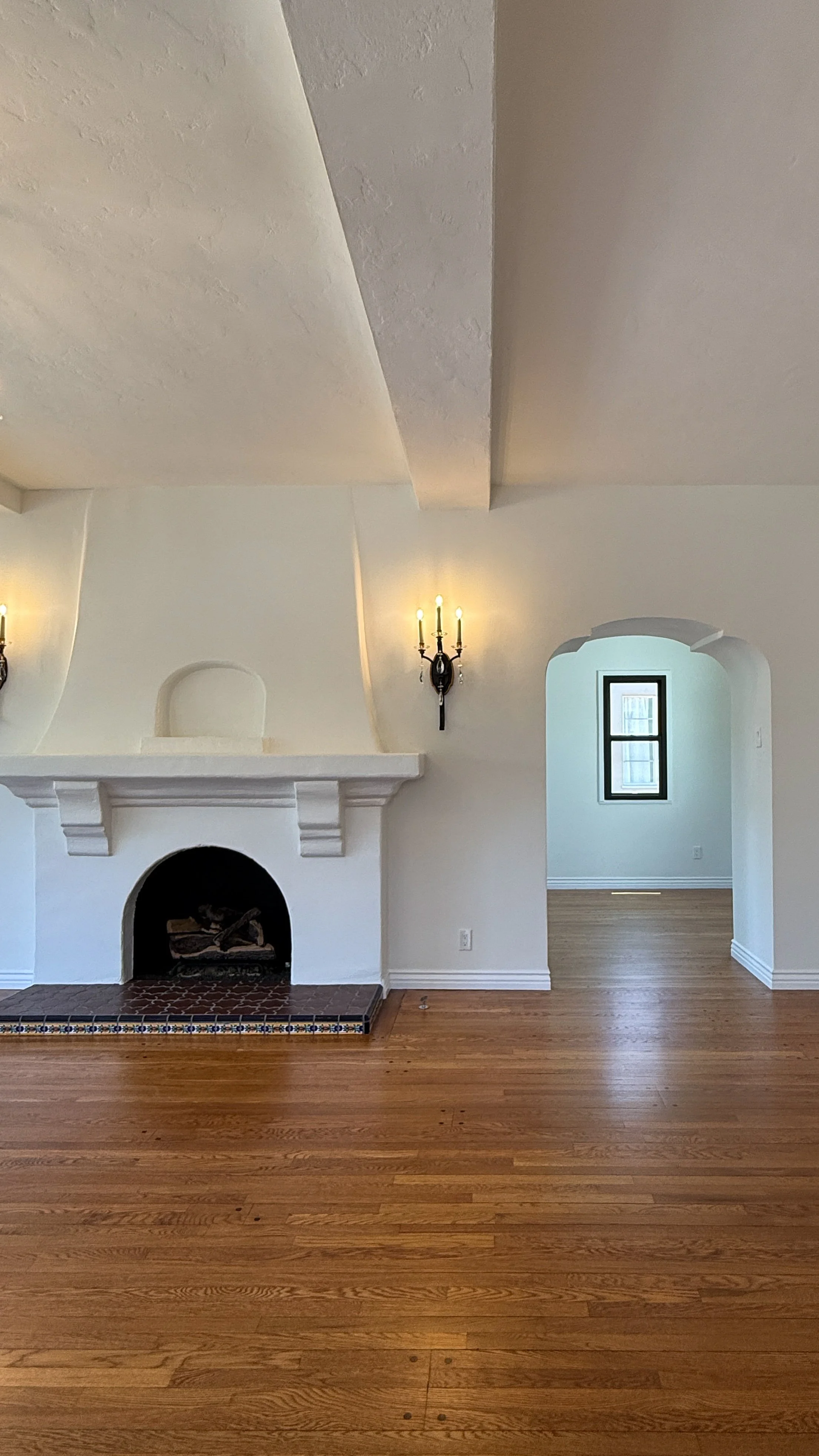 Interior view of a room with a white fireplace, wooden floors, a window with black trim, and a doorway with an arched top. There are two wall-mounted light fixtures on either side of the fireplace.