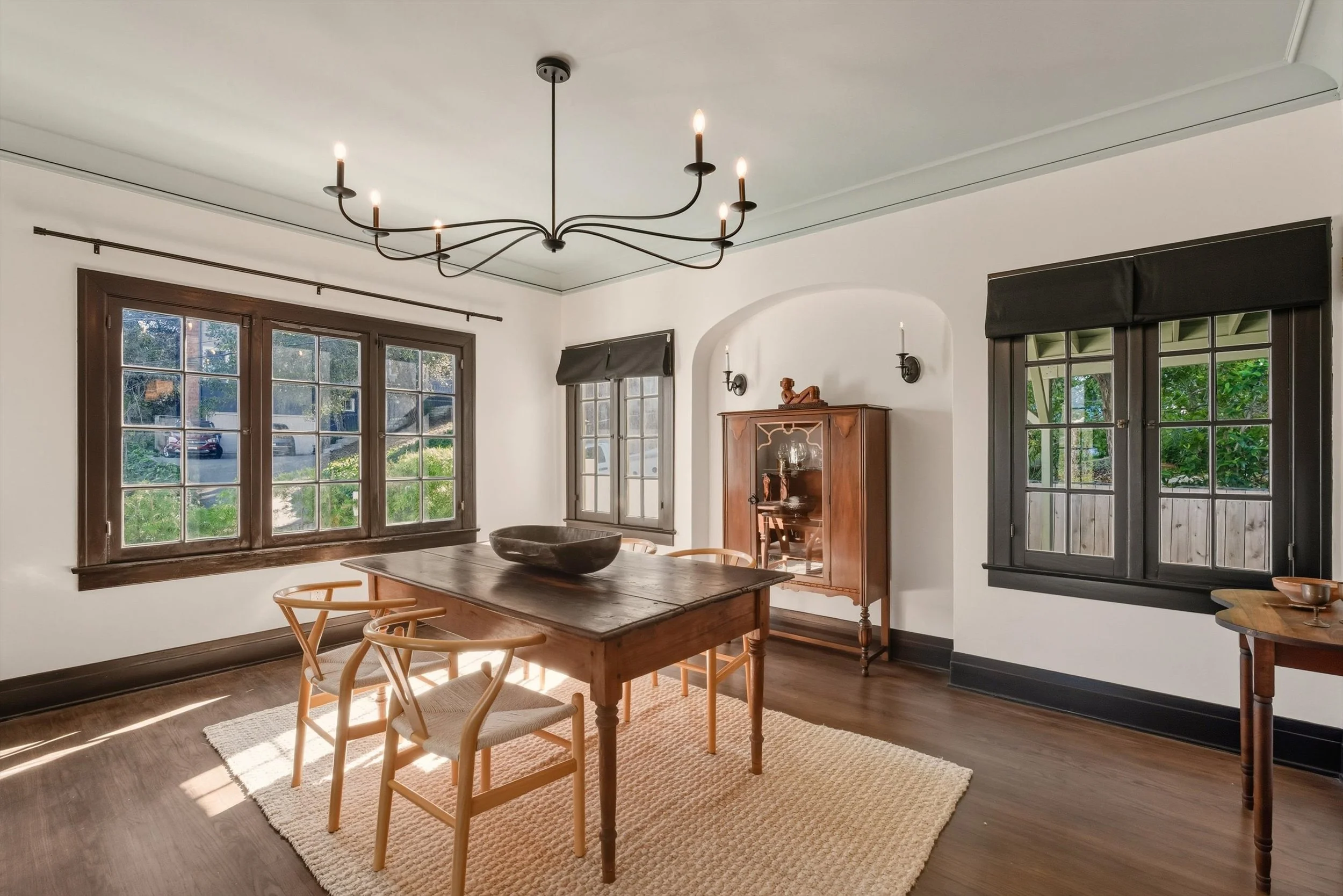 A dining room with wooden table and chairs, large windows, black window frames, a vintage wooden cabinet with a decorative sculpture on top, and black wall-mounted candle holders. Sunlight streams through the windows.