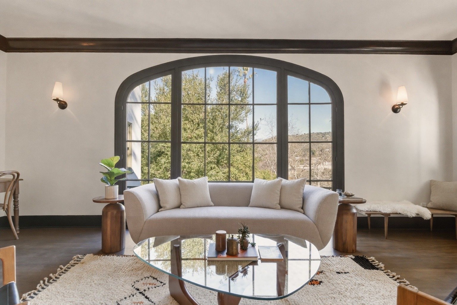 Living room with a large arched window, white sofa with cushions, wooden side tables, potted plant, glass coffee table, patterned rug, wall sconces, and a bench with a pillow