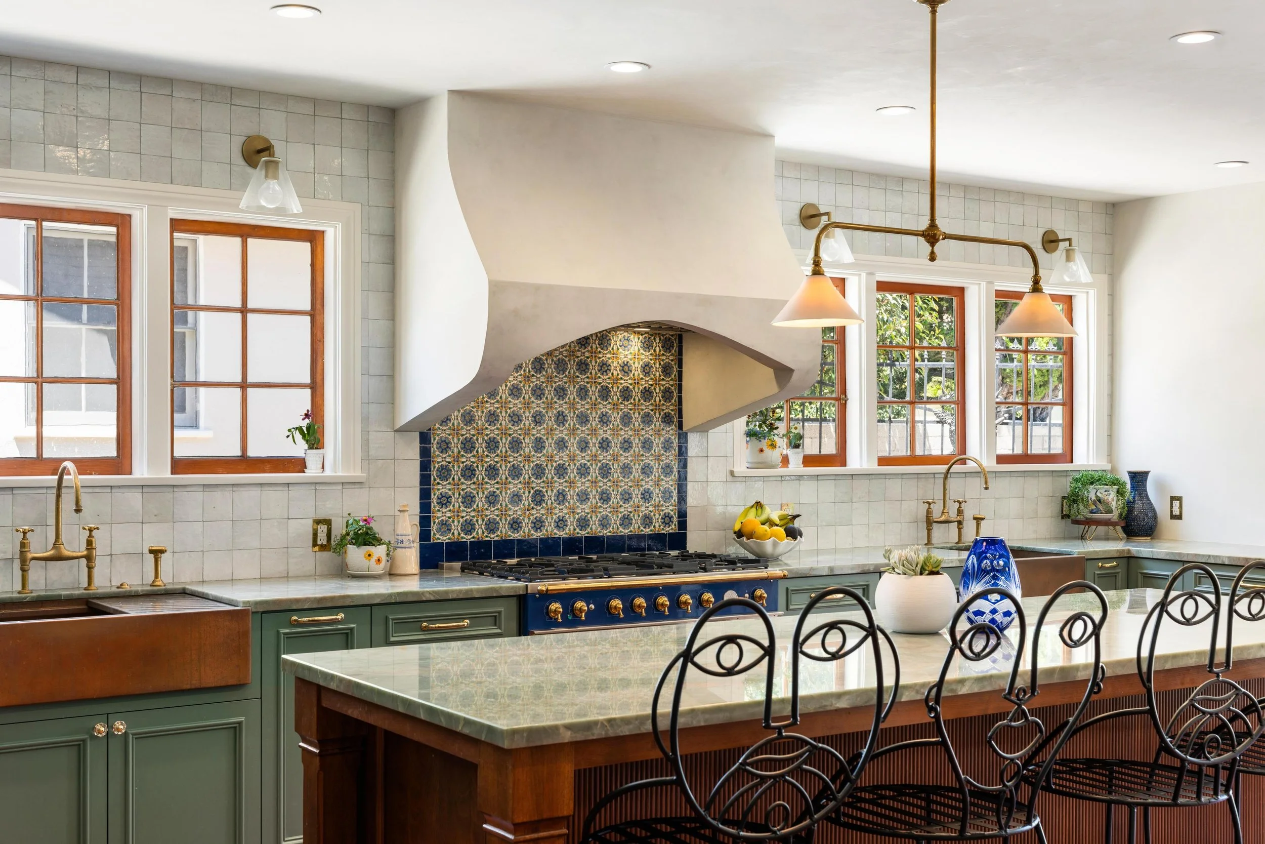 A bright, spacious kitchen with green cabinets, a large island with a marble countertop, and multiple windows letting in natural light. There is a decorative fireplace with patterned tiles and various kitchen accessories and plants on the countertops.