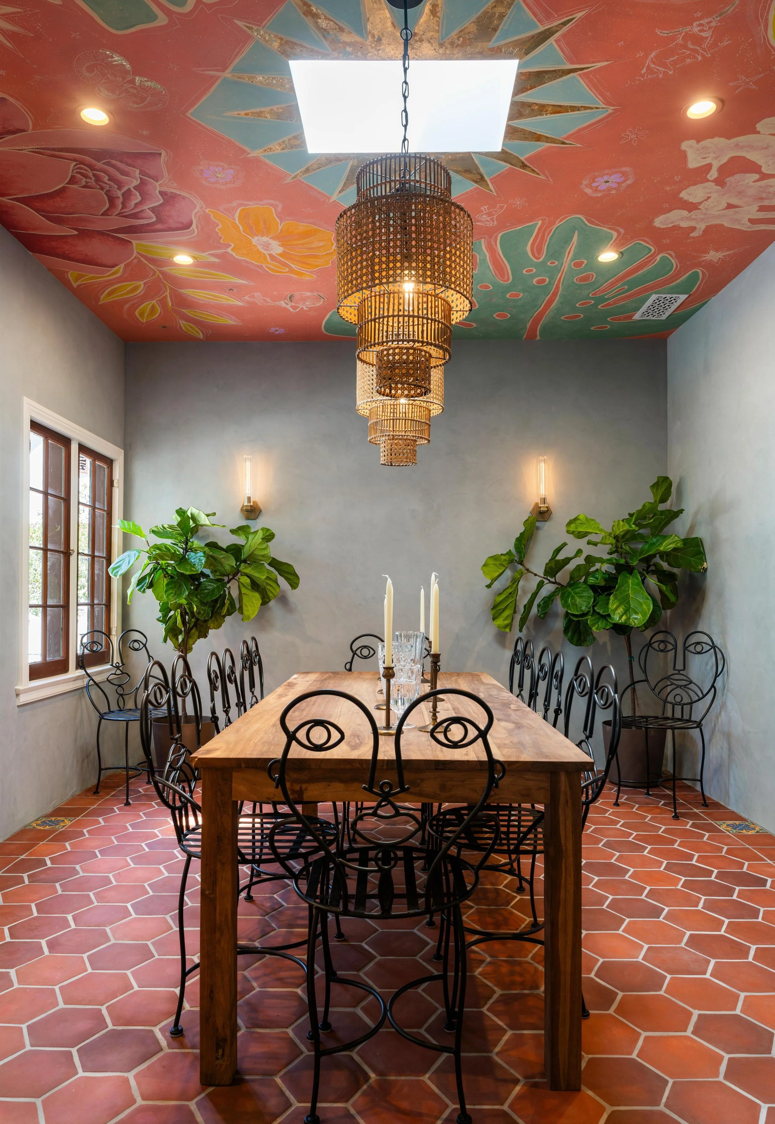 A dining room with a wooden table, black metal chairs, large green leafy plants, terracotta tile floor, two wall sconces, a window, and a colorful painted ceiling with floral and animal designs, and a hanging wicker chandelier.