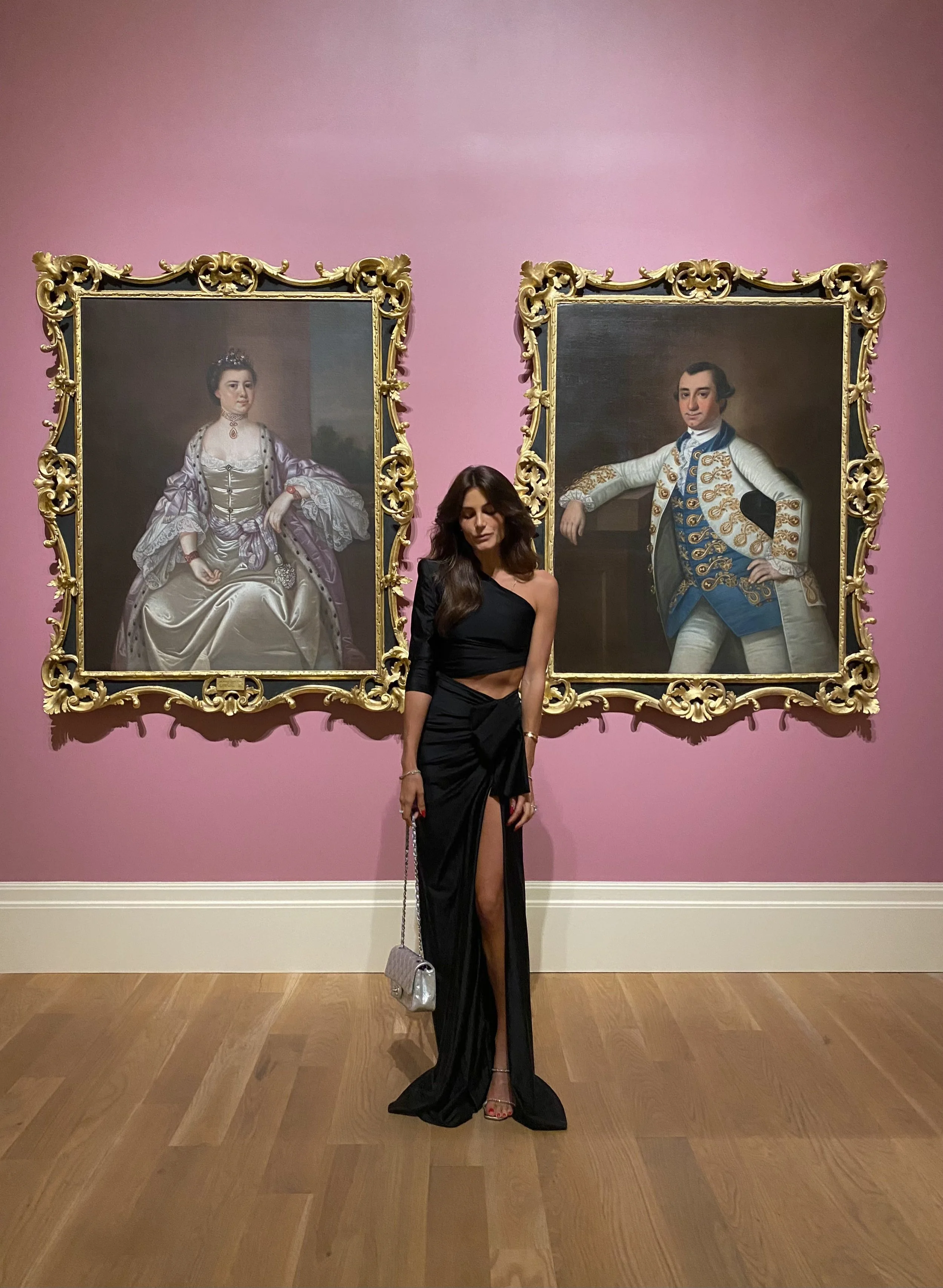 Woman with long dark hair wearing a black dress with a thigh-high slit, standing in front of two large framed portrait paintings on a pink wall in an art gallery.