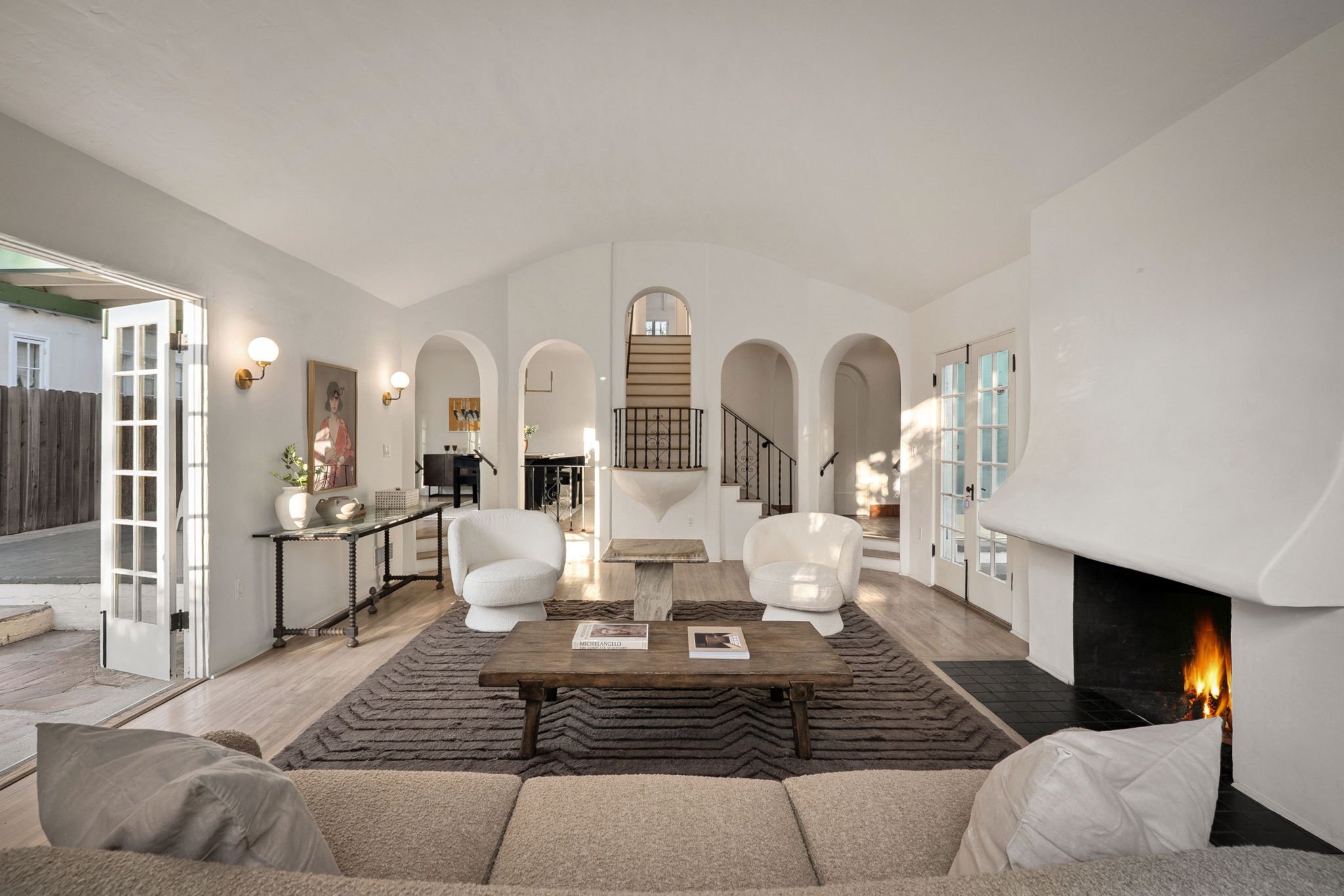 Living room with white walls, hardwood flooring, two white armchairs, a wooden coffee table, a fireplace with a visible fire, and large glass doors allowing natural light inside.