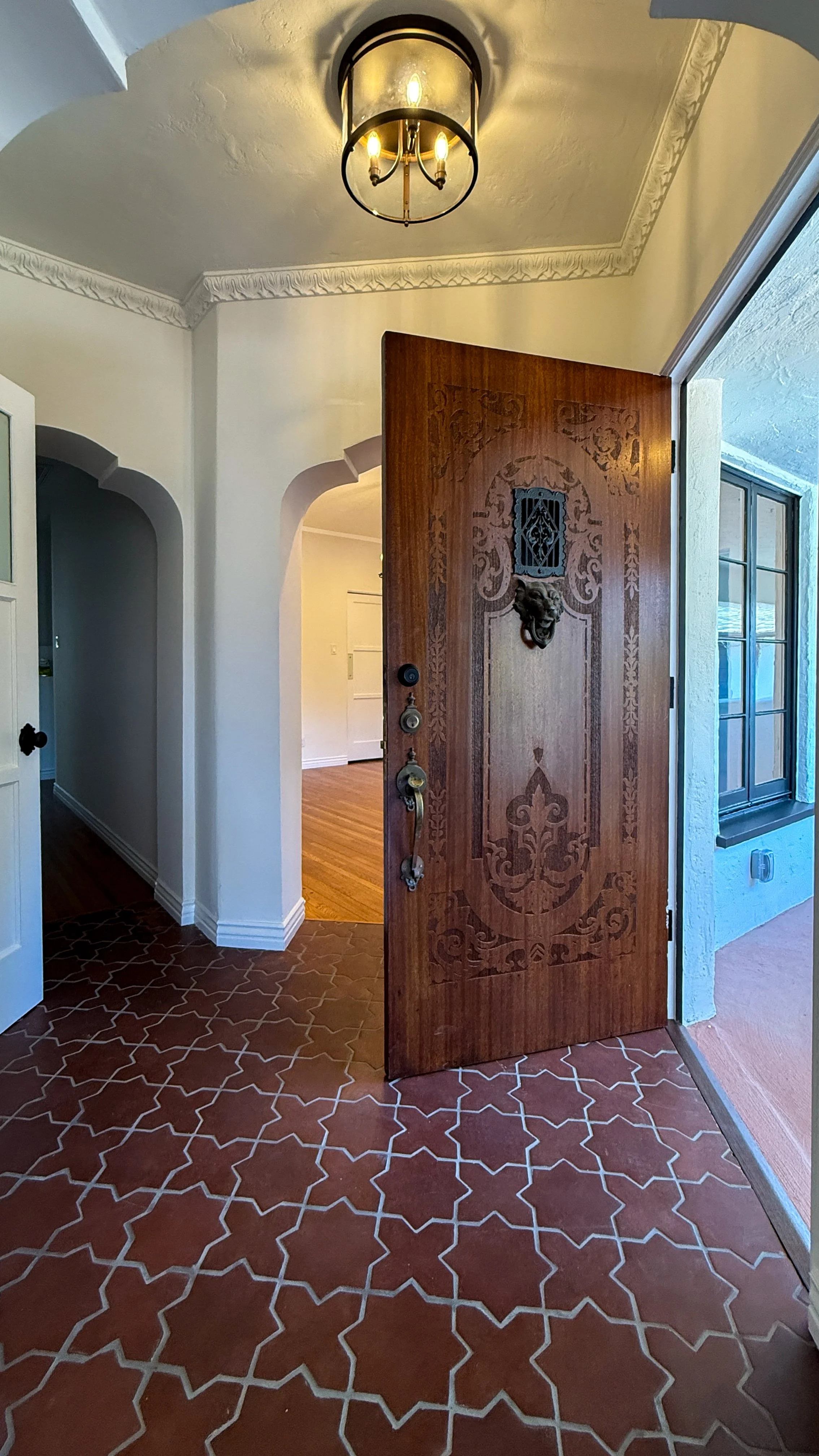 Entrance with a wooden door carved with intricate designs and a lion head door knocker, open to reveal a hallway with wooden flooring, a nearby window, and a ceiling light fixture.