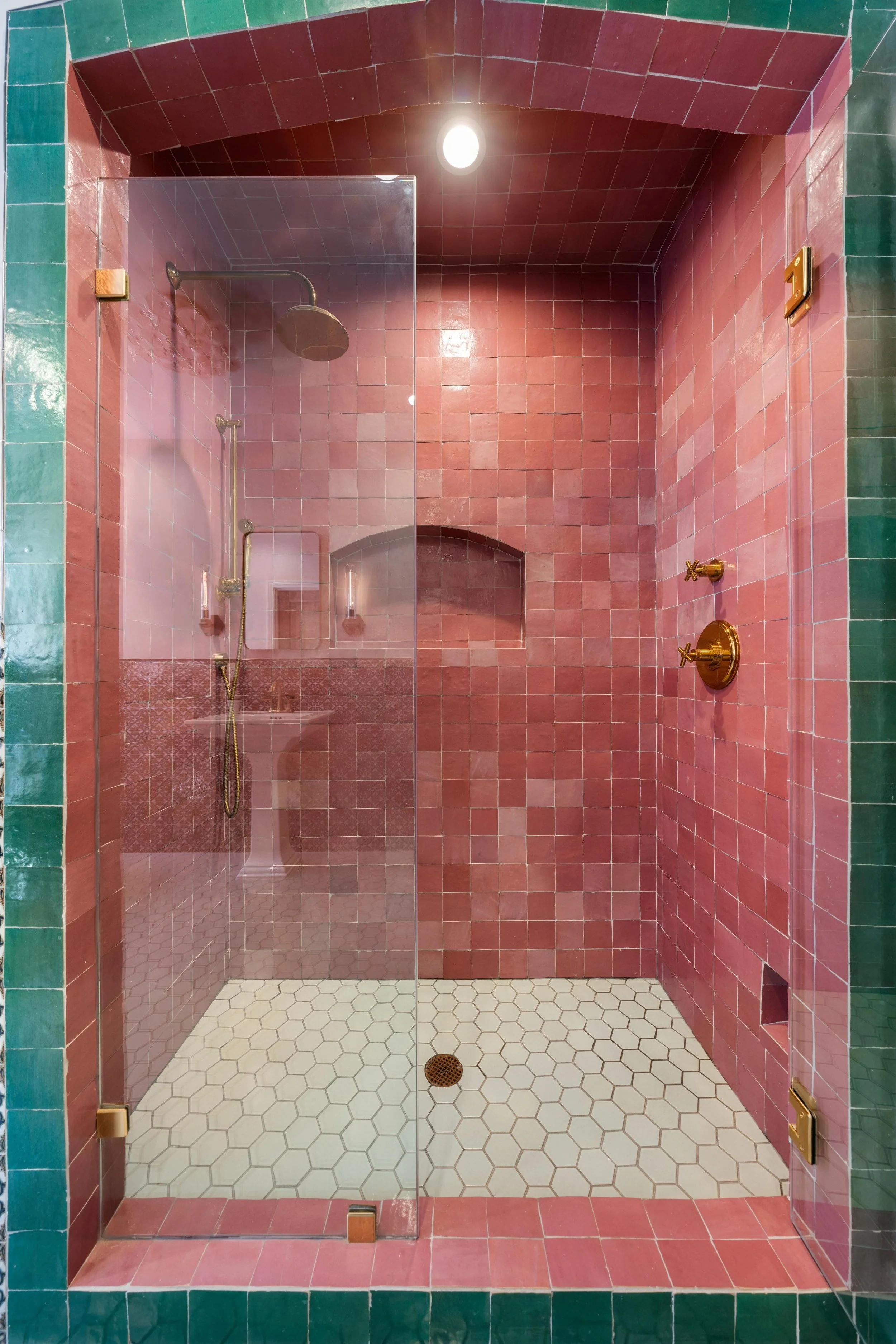 A bathroom shower with pink tiles on the walls and white hexagonal tiles on the floor, separated by a glass partition, with brass fixtures, a built-in niche, and a ceiling light.