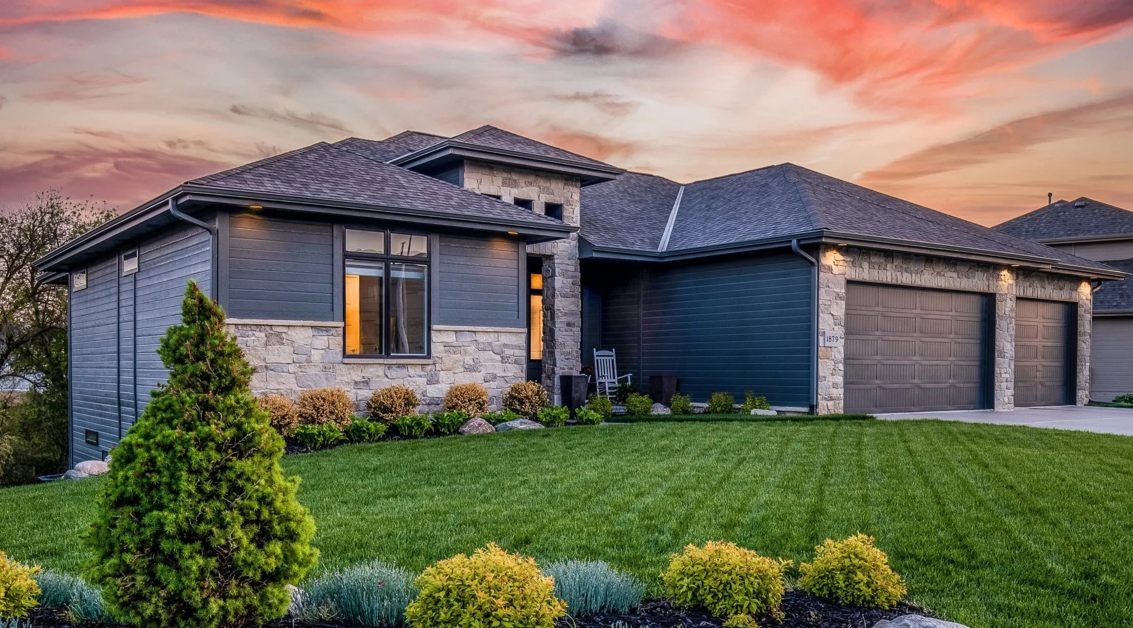 A modern house with dark gray siding and stone accents, a two-car garage, a well-maintained green lawn, and landscaped plants in front, with a colorful sunset sky in the background.