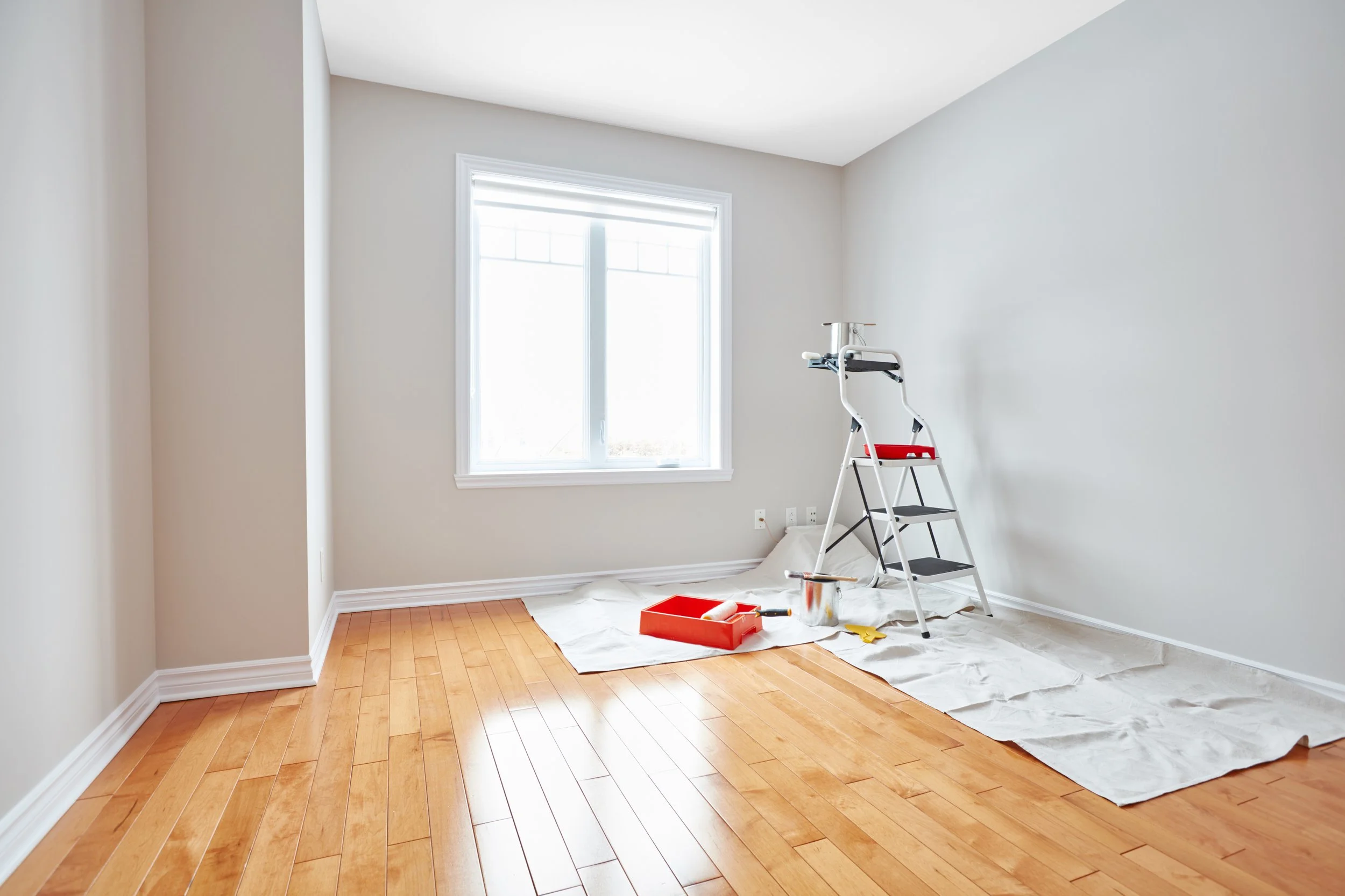 A room being painted or renovated with painting supplies on the floor, including a paint roller and tray, a ladder, and drop cloths, with a window letting in natural light.