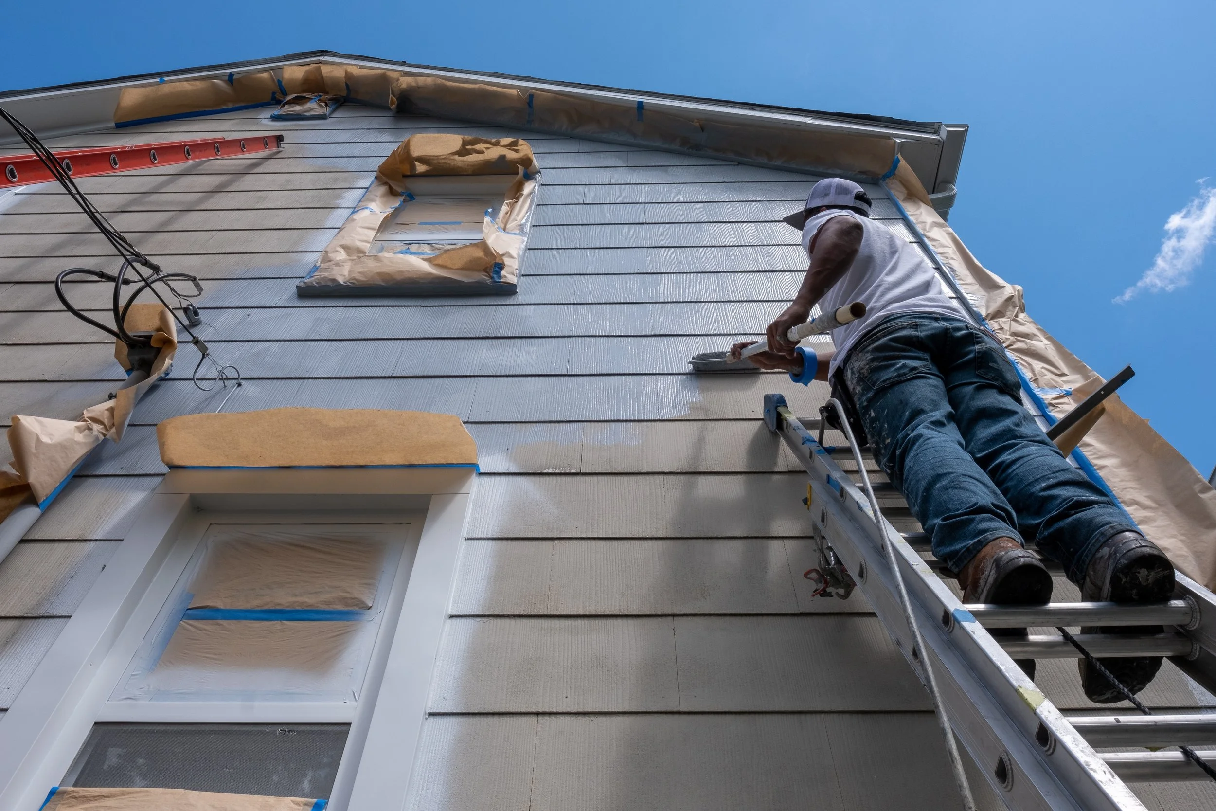 A worker on a ladder painting the exterior wall of a house. The wall is beige, and the worker is painting with white. The worker is wearing a white shirt, jeans, a cap, and is using a paint roller. The house has a window below the area being painted, and the sky is blue with a few clouds.