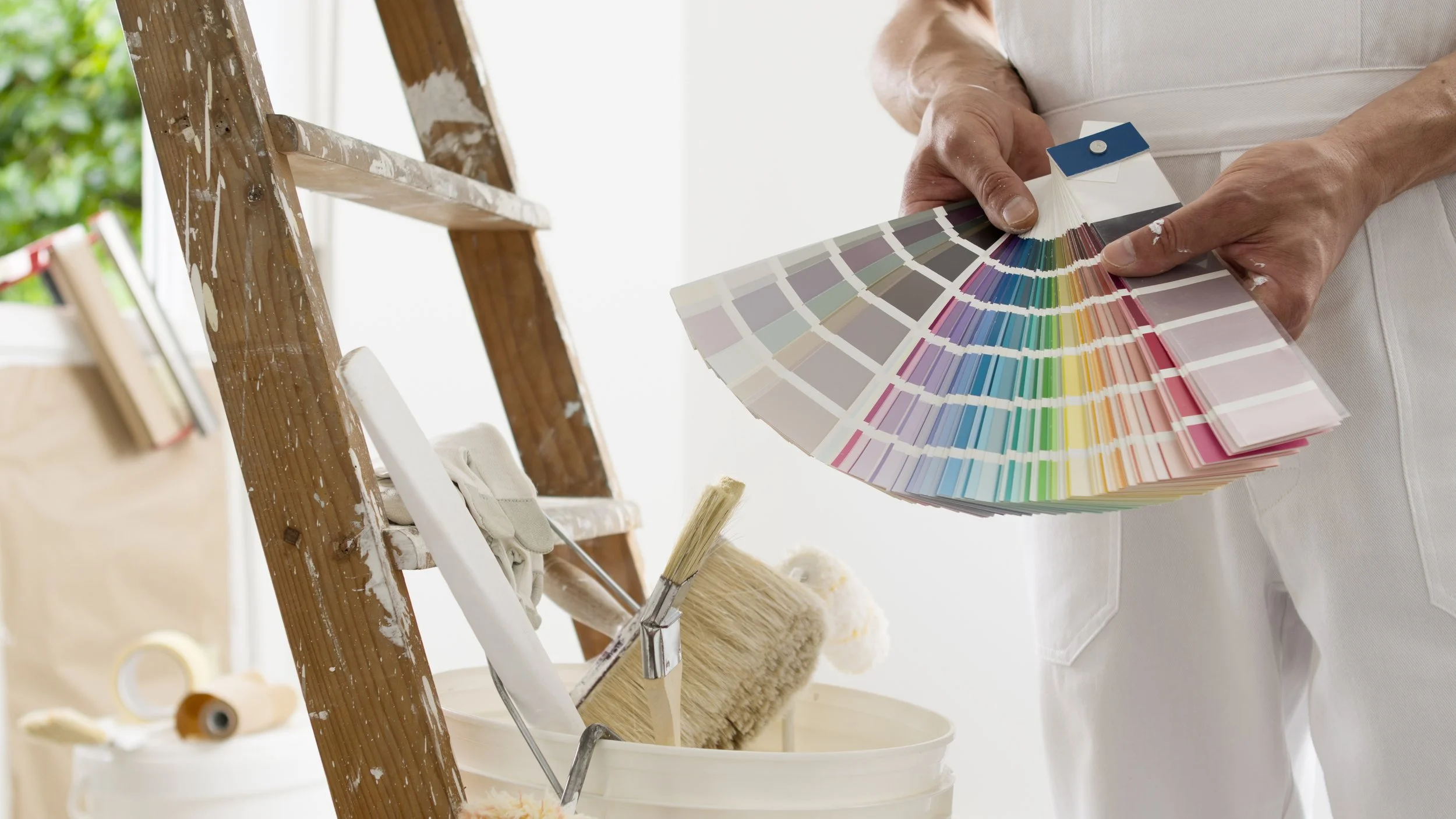A person holding a fan deck of paint color swatches next to a wooden ladder with paint splatters, paintbrushes, and painting supplies in the background.