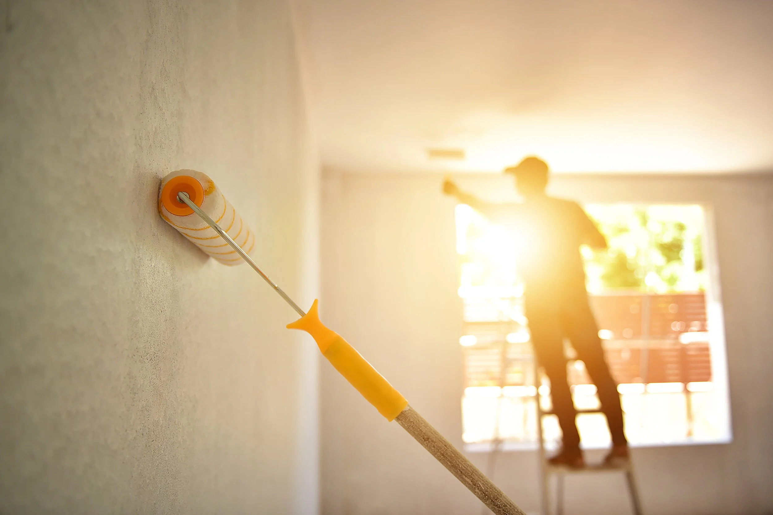 A close-up of a paint roller with orange and white striped pattern and yellow handle, leaning against a wall. The background shows a bright room with sunlight and two people in silhouette, one holding a drink, near an open door leading outside.