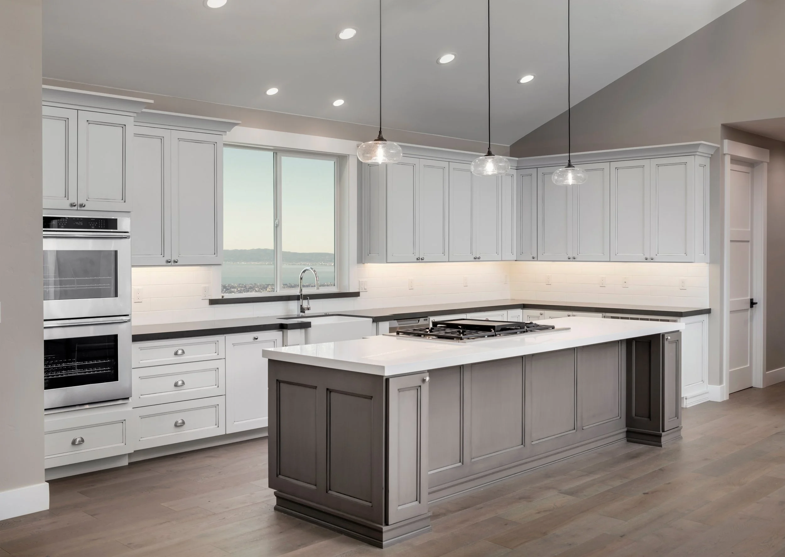 Modern kitchen with painted, refinished, white cabinets, a gray island, stainless steel oven, and pendant lighting, large window overlooking a scenic view.