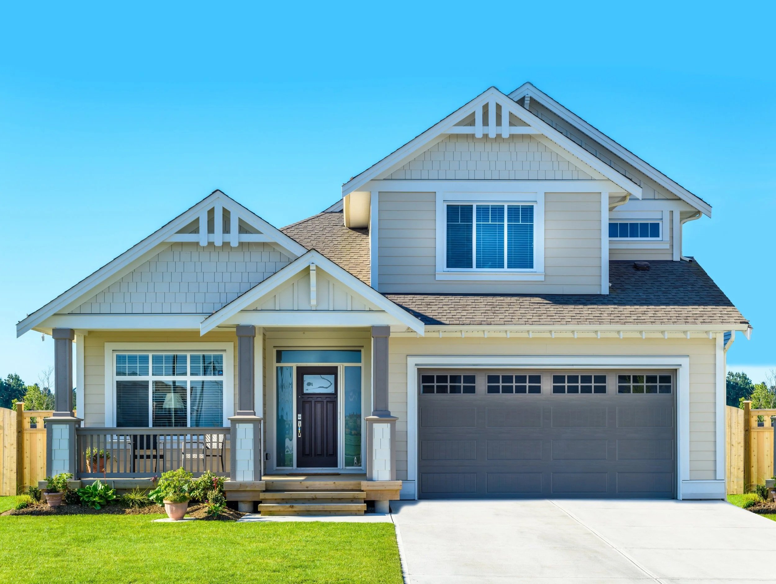 Front view of a two-story house with painted beige siding, white trim, large windows, a small front porch with stairs, a garage on the right, a well-maintained lawn, and a clear blue sky in the background.