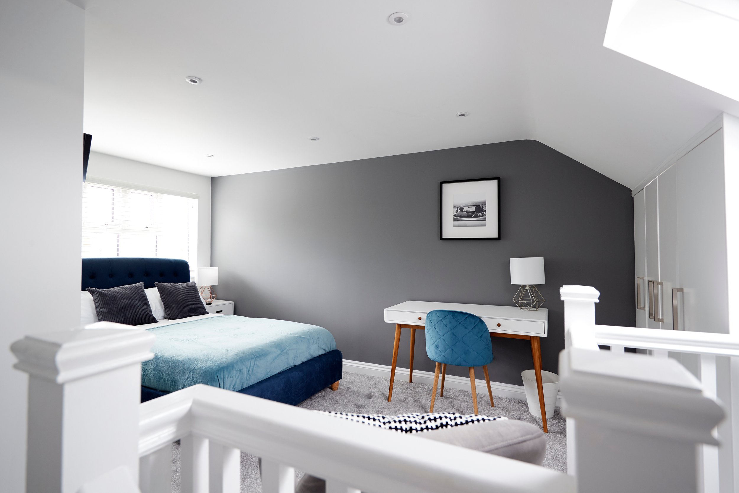 A modern bedroom with a blue bed, black pillows, white nightstands with lamps, a gray accent wall with a black-and-white picture, a white desk with a blue chair, a window with white blinds, and a gray carpet, seen through a white railing.