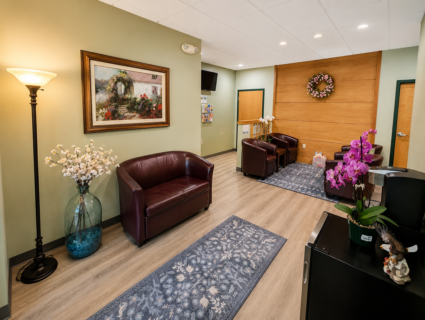 Waiting room with green walls and wooden panel accent wall, featuring leather armchairs and a sofa, framed landscape painting, flowers in vases, wall-mounted TV, and area rugs.