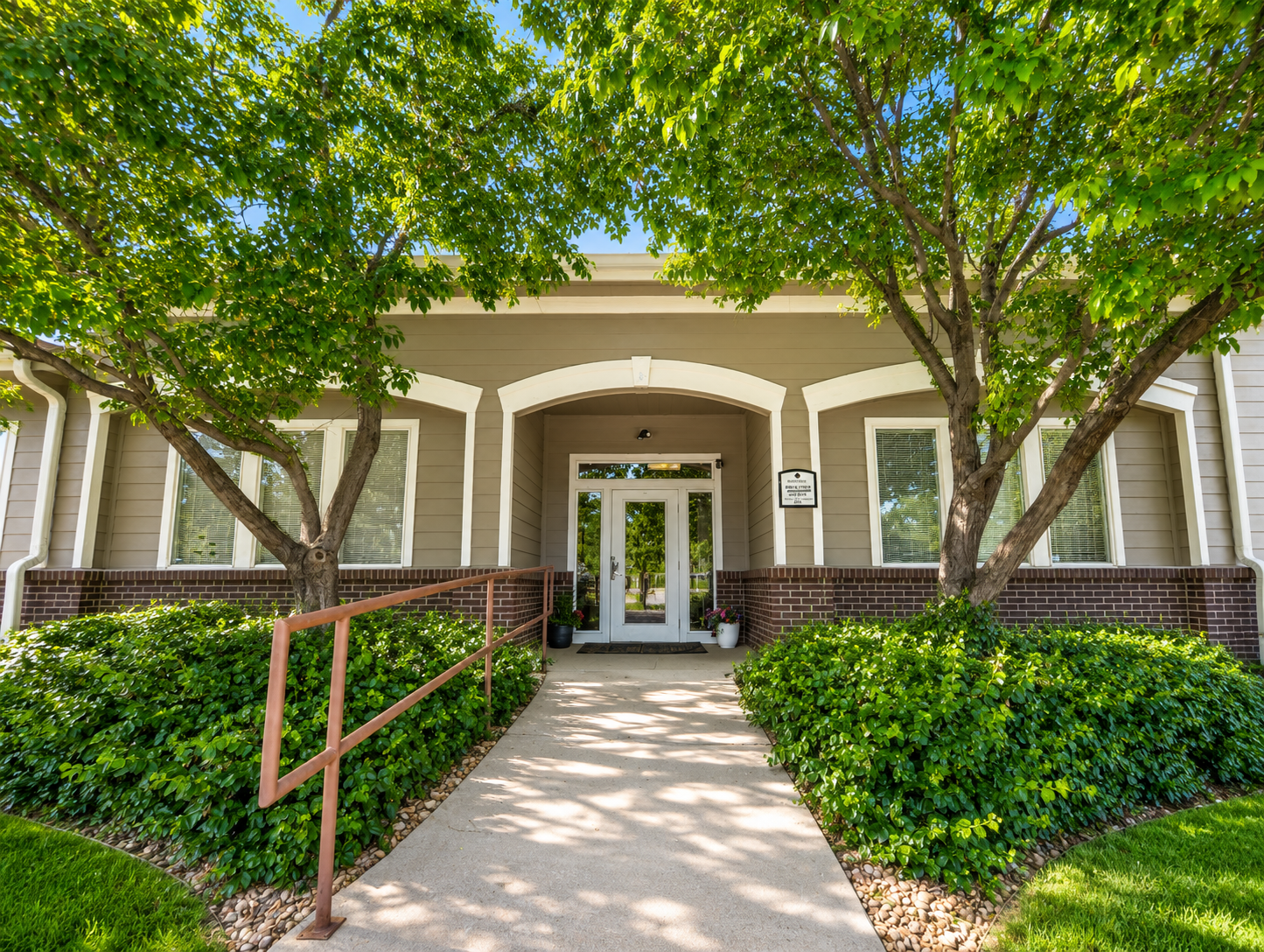 Entrance to Orchid dental with a glass door, surrounded by trees and well-maintained bushes, with a paved walkway and a pink handrail leading to the door.