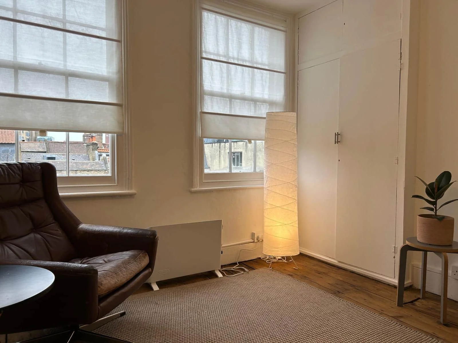 Interior shot of Calm Harbour Therapy room, featuring brown leather armchair, a floor lamp with warm light, large windows with white blinds and hardwood floors.