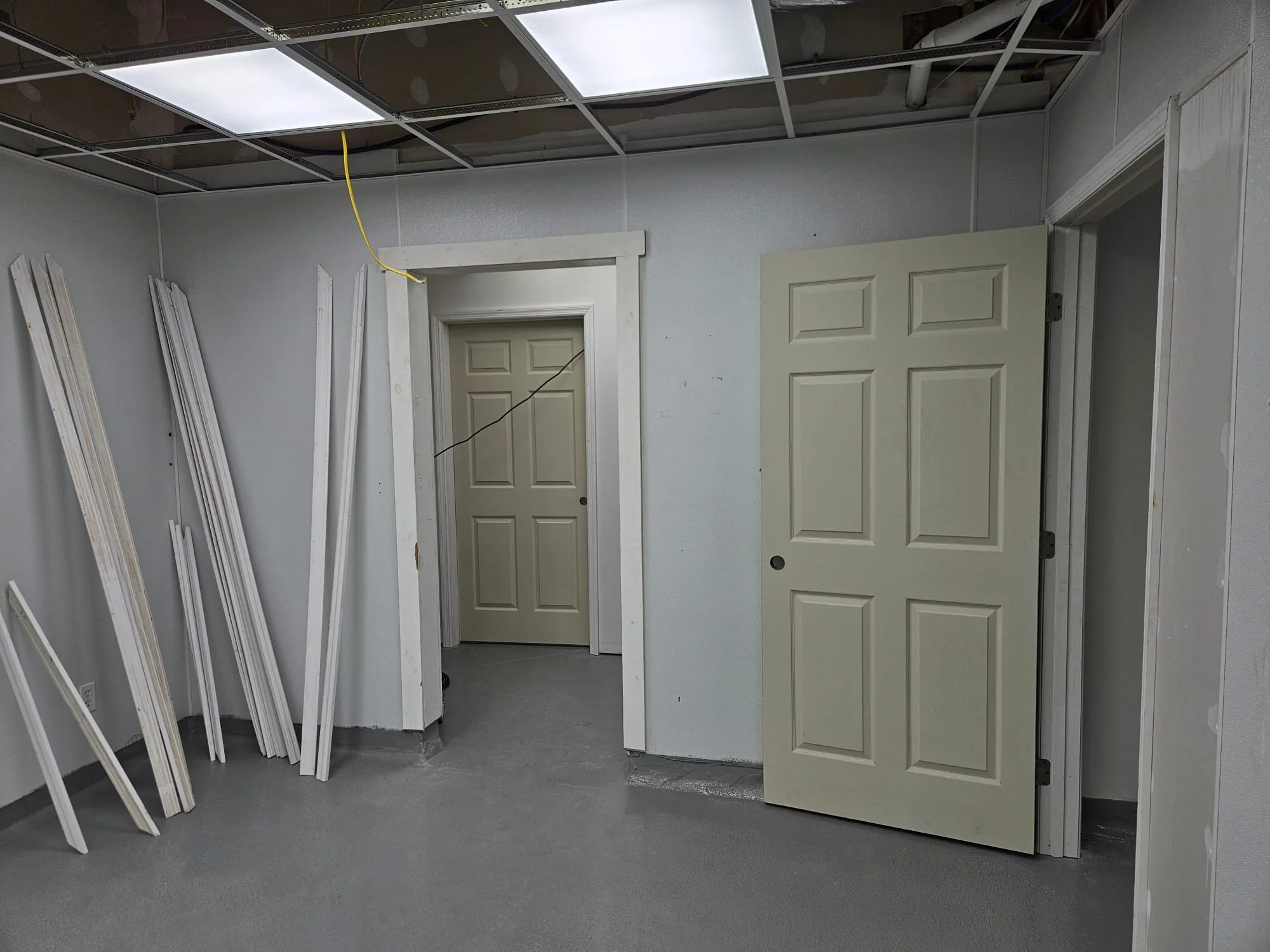 A room with a ceiling with partially removed ceiling tiles, some hanging wires, an open door, and several white wooden frames leaning against the wall.