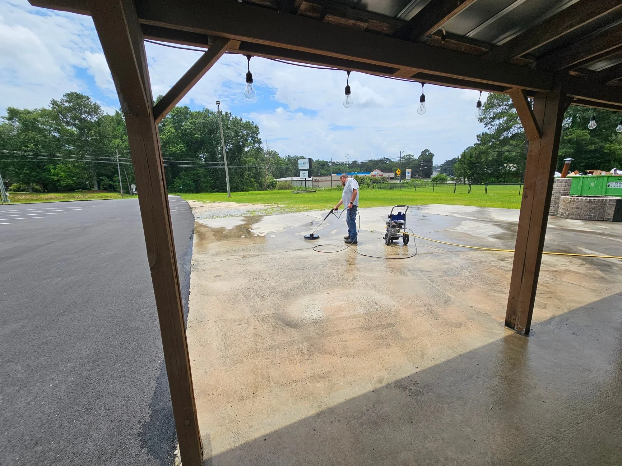 A man pressure washing a concrete surface outside a building. He is wearing a white shirt and jeans, with a portable pressure washer connected to a yellow hose. The scene is viewed from under a wooden structure with hanging light bulbs, with a parking lot and grassy area in the background.