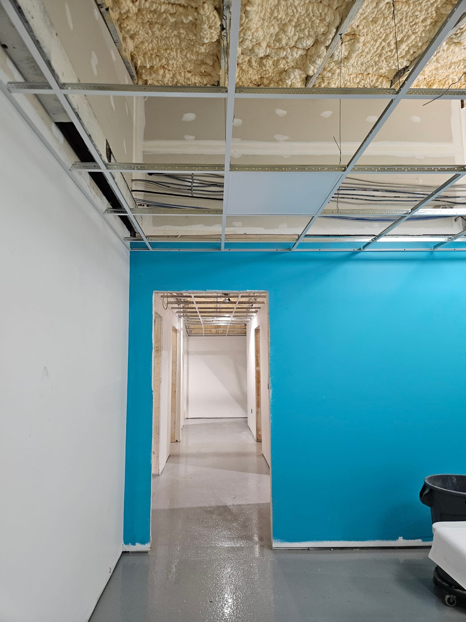 Construction site with drywall partially installed and ceiling insulation exposed in an interior hallway, blue wall on the right and doorway leading to another room in the background.