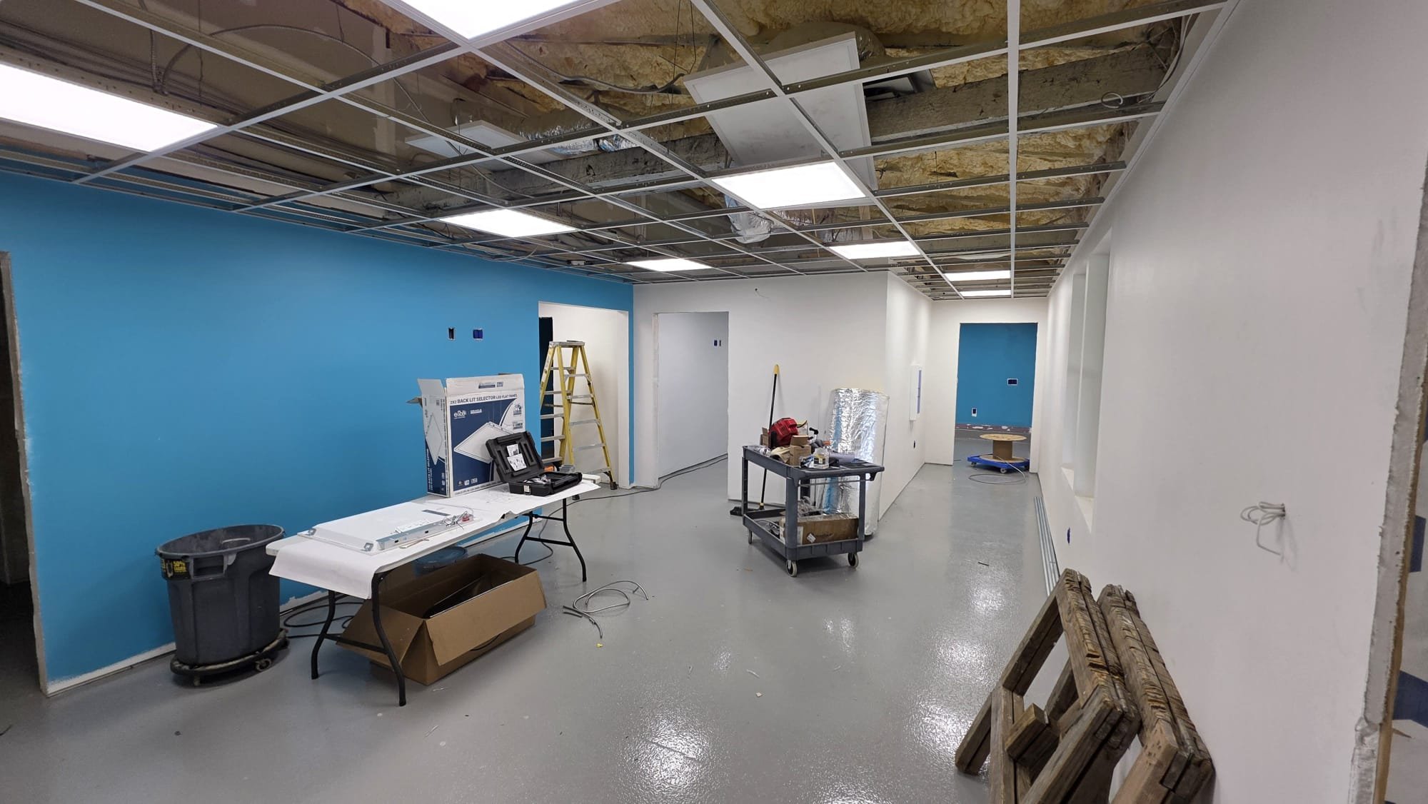 Interior of a construction or renovation site with partial ceiling exposed, showing insulation and metal framing. The walls are painted in light and dark blue, and there are construction tools, a ladder, a worktable with equipment, a trash bin, and wooden pallets on the floor.