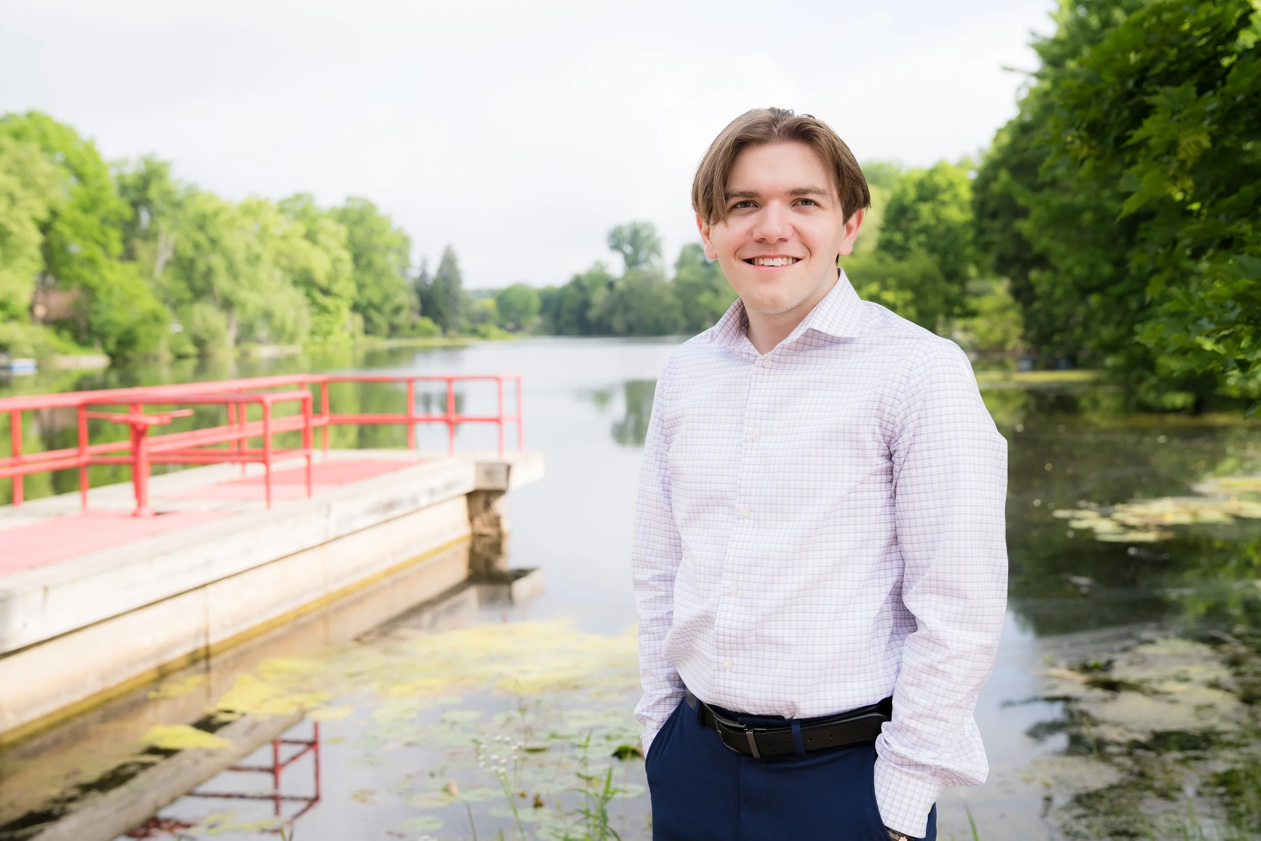 A young man smiling outdoors near a body of water with lush green trees in the background.
