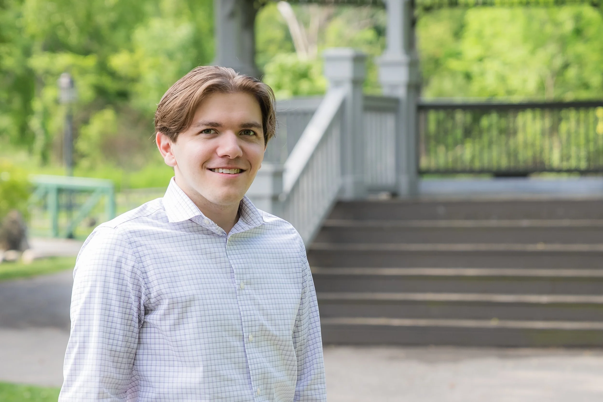 Young man with brown hair wearing a white checkered shirt standing outdoors near a wooden staircase and green trees.