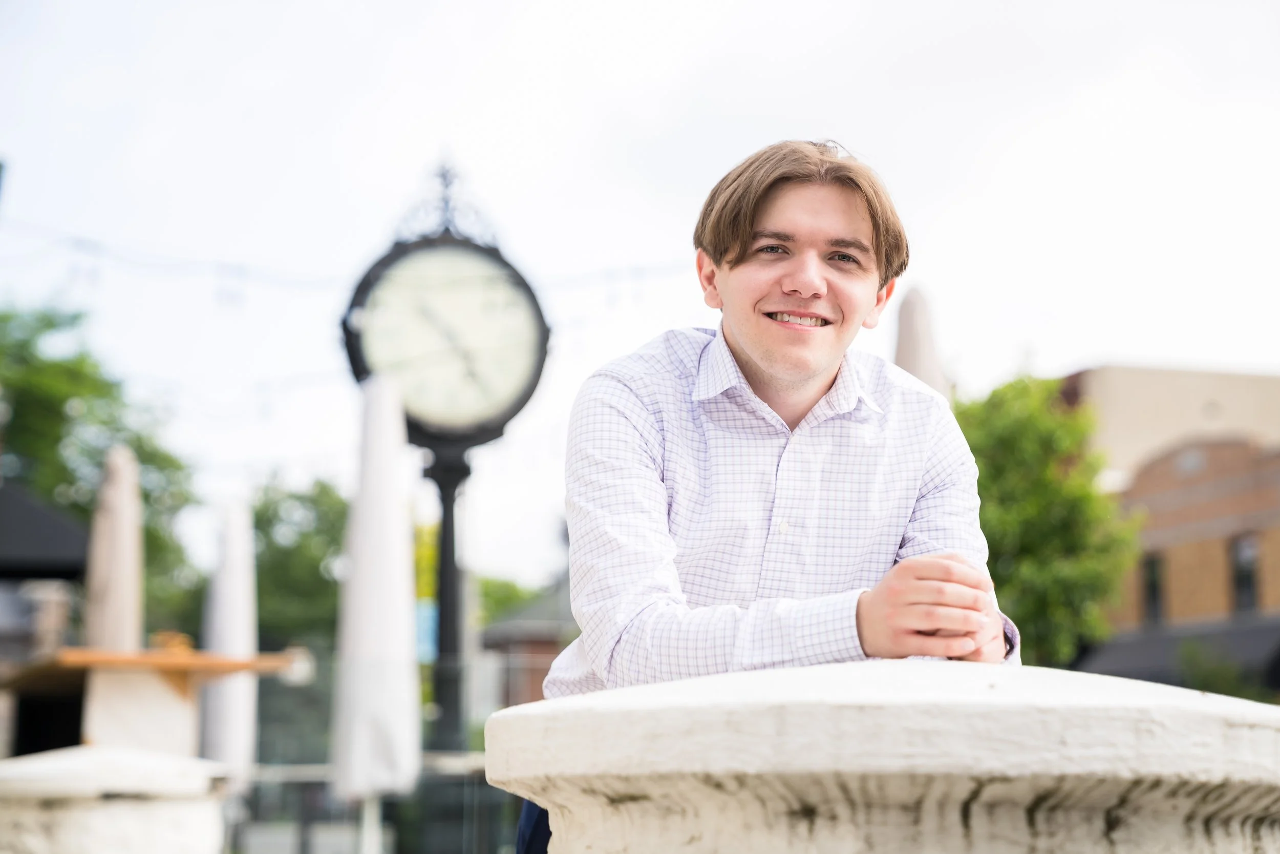Young man with light brown hair smiling, sitting outdoors at a round stone table with a blurred clock and trees in the background.