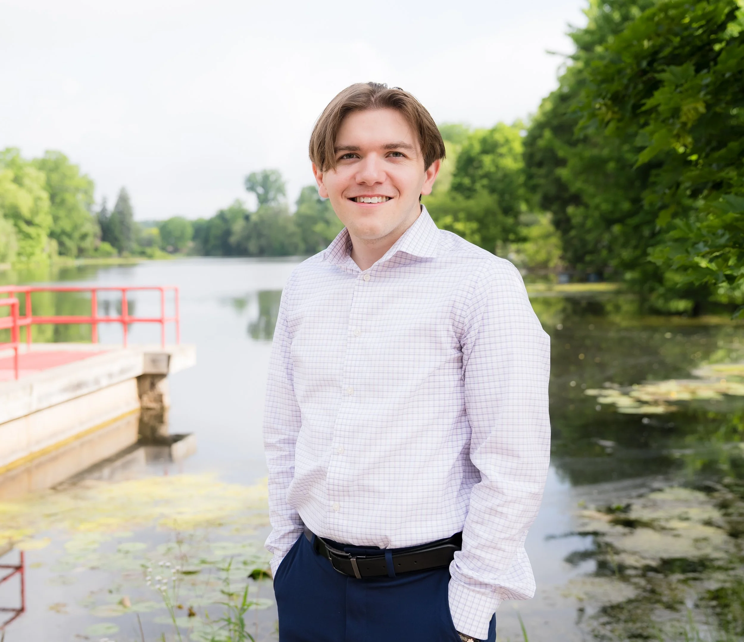 A young man with brown hair, wearing a light purple checkered shirt and dark pants, stands outdoors near a lake with green trees in the background, smiling at the camera.
