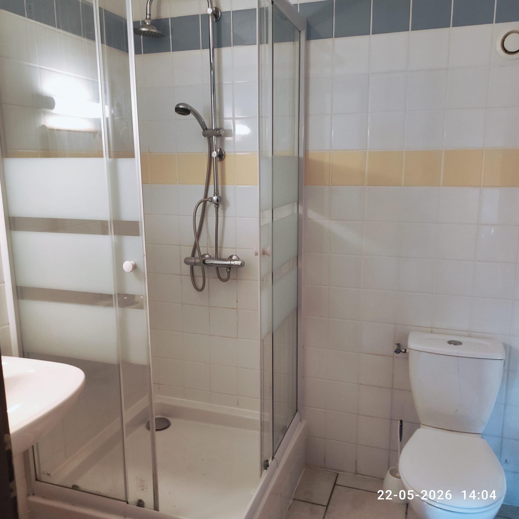 Een badkamer met een inloopdouche, een toilet en een wastafel, betegeld in wit, met een horizontale lijn van beige tegels. Er is een sketch onder de douche en een klok op de muur. De foto is genomen op 22 mei 2026 om 14:04.