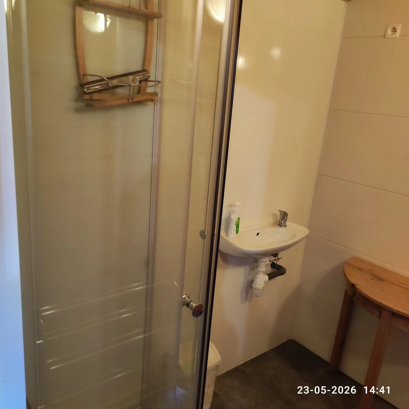 Een kleine badkamer met een douchecabine, een wastafel en een houten krukje. Op de muur hangt een spiegel en een muurcontact. De vloer is bedekt met donkere tegels en de muur is licht van kleur.
