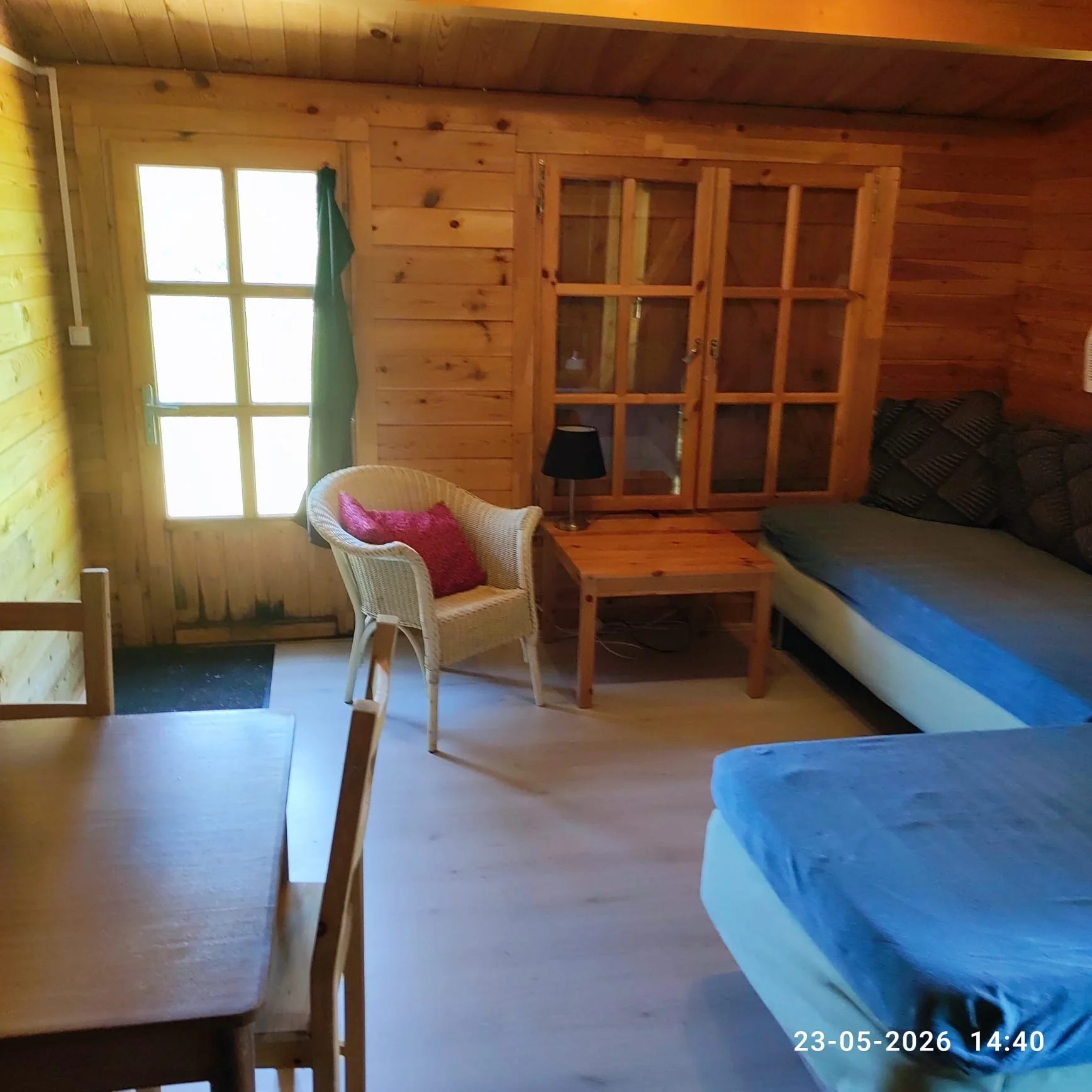 Interieur van een houten huis met een raam, een deur met glasramen, een witte stoel met een roze kussen, een tafel, een bank met kussens en een lamp, met een houten muur en plafond.