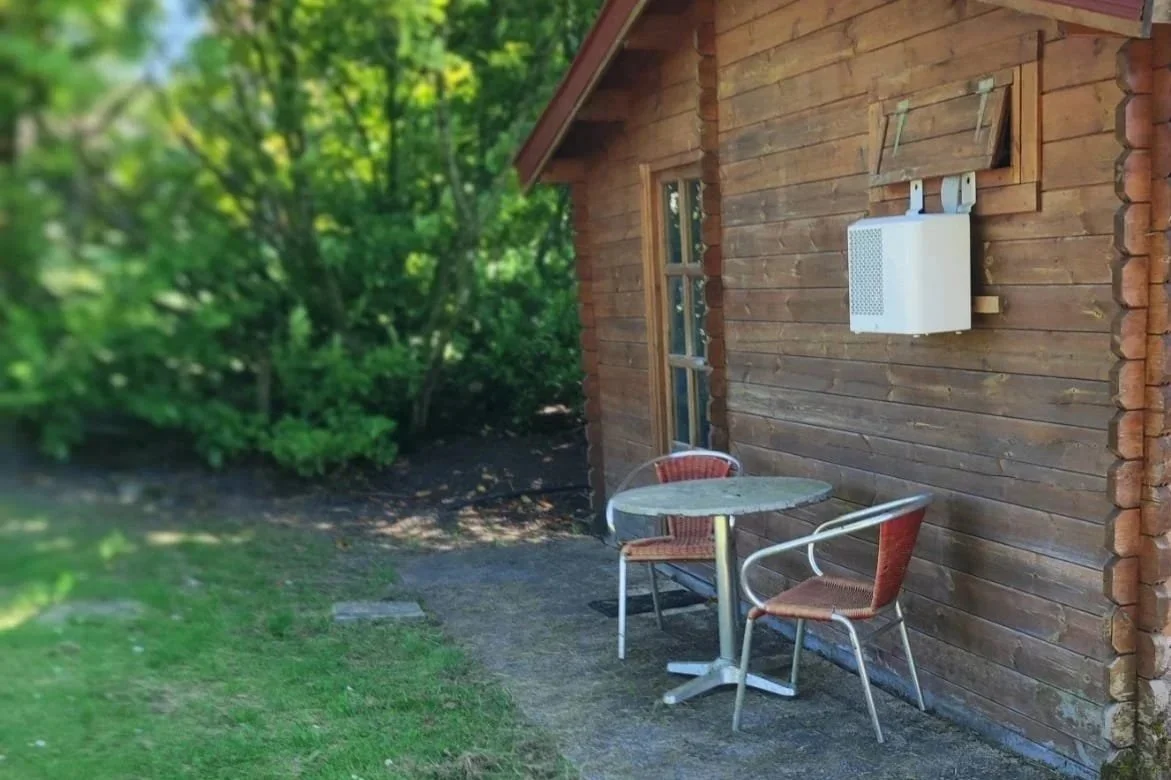 Buitenzitje met tafel en twee stoelen naast een houten huis, omgeven door groen bomen.