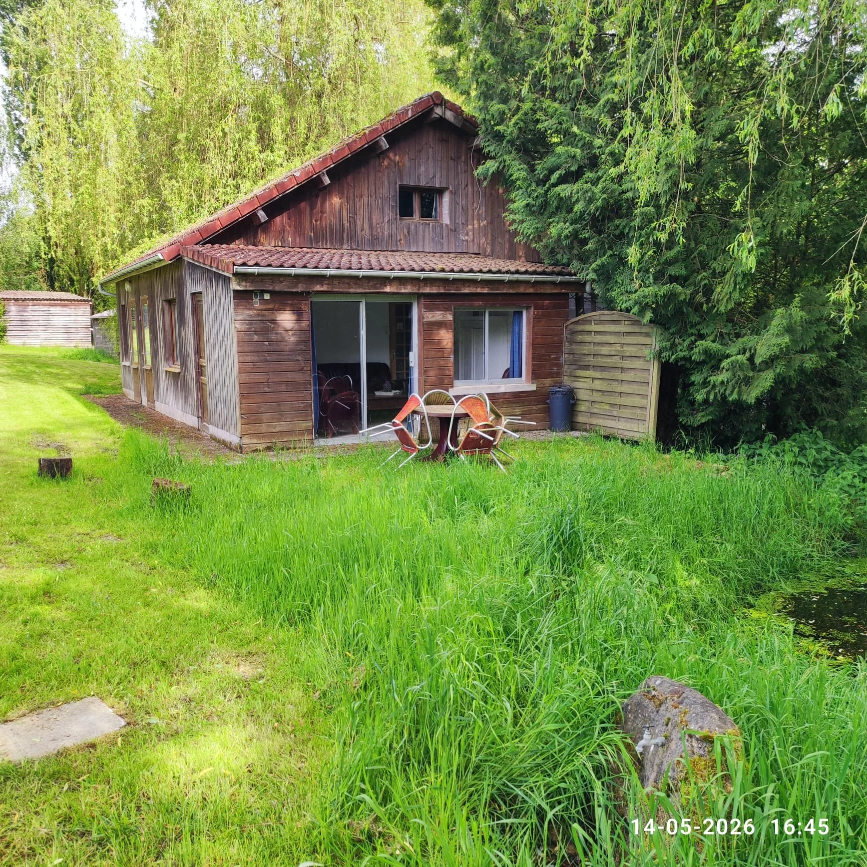 Een houten huis in een groene tuin met een gazon en een vijver. Op de foto staat een tafel en vier stoelen. In de achtergrond zijn bomen en struiken te zien. Bovenaan de afbeelding bevindt zich de datum en tijd: 14-05-2026 16:45.