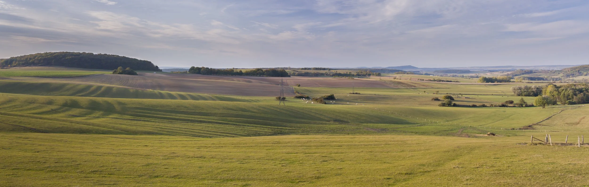 Uitzicht op uitgestrekte groene en bruine landbouwvelden, heuvels en een blauwe hemel met wat wolken in het landschap.