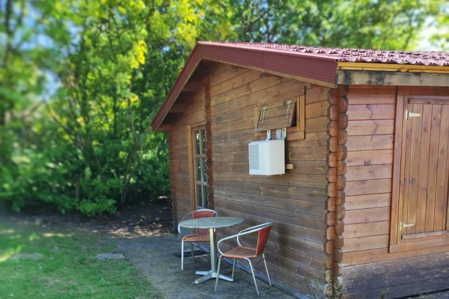 Een houten schuur met een rode dak en een klein terras met twee rode buitenstoelen en een ronde tafel, omringd door groene bomen.