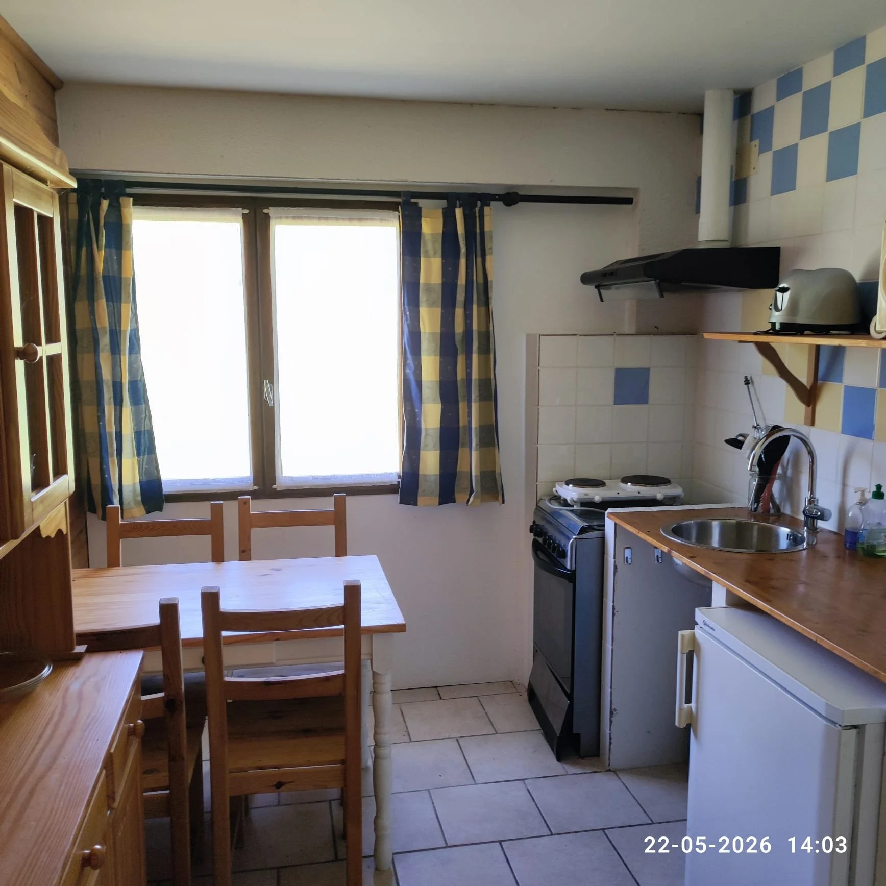 De afbeelding toont een keuken met een houten eettafel en vier houten stoelen, een raam met blauwe en gele geruite gordijnen, een gasfornuis, een wasmachine, een muur met blauwe en witte tegels, een afzuigkap, een broodrooster en een gootsteen. Op de